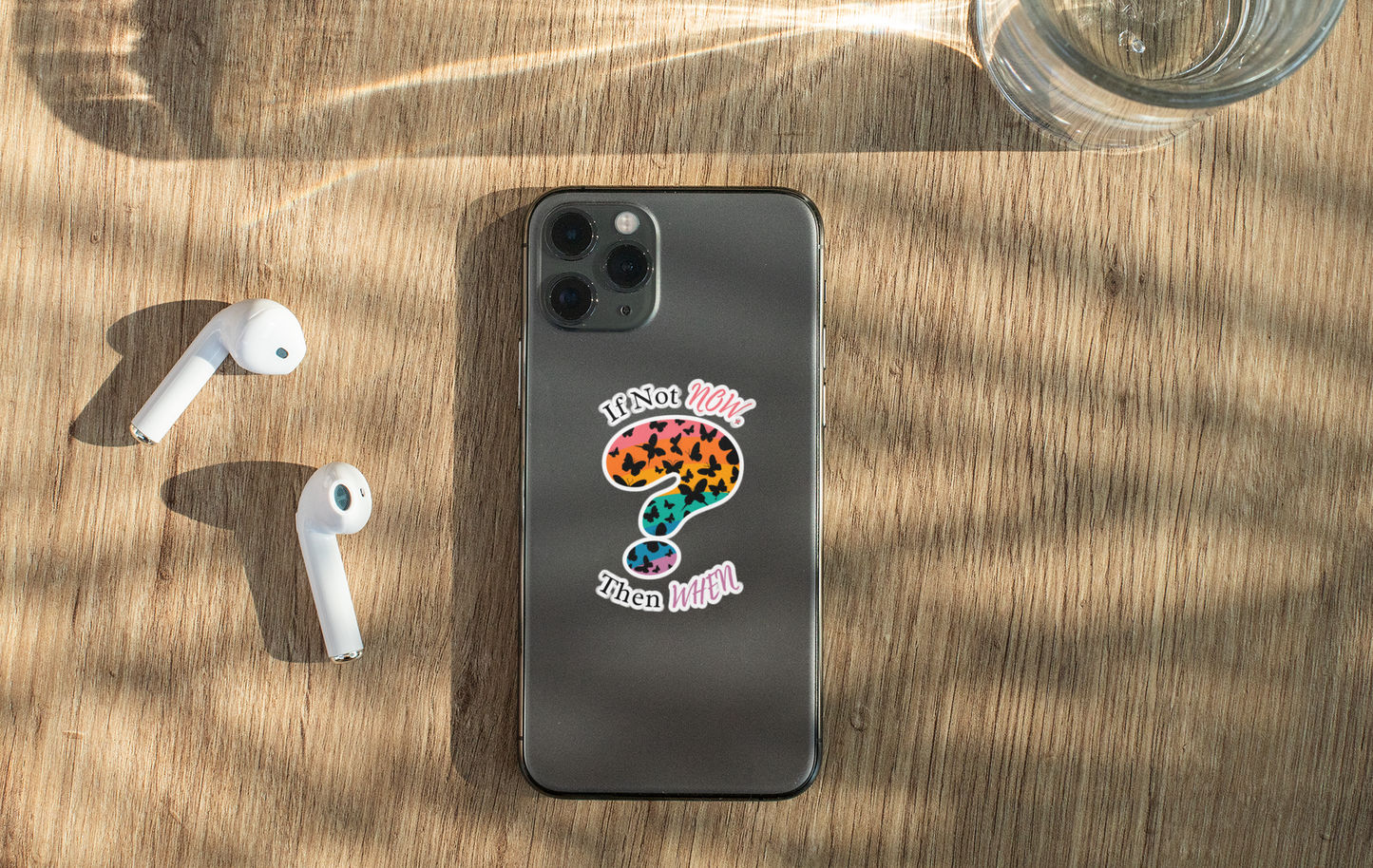 phone with “if not now then when” motivational vinyl sticker, colorful case and airpods on wooden surface
