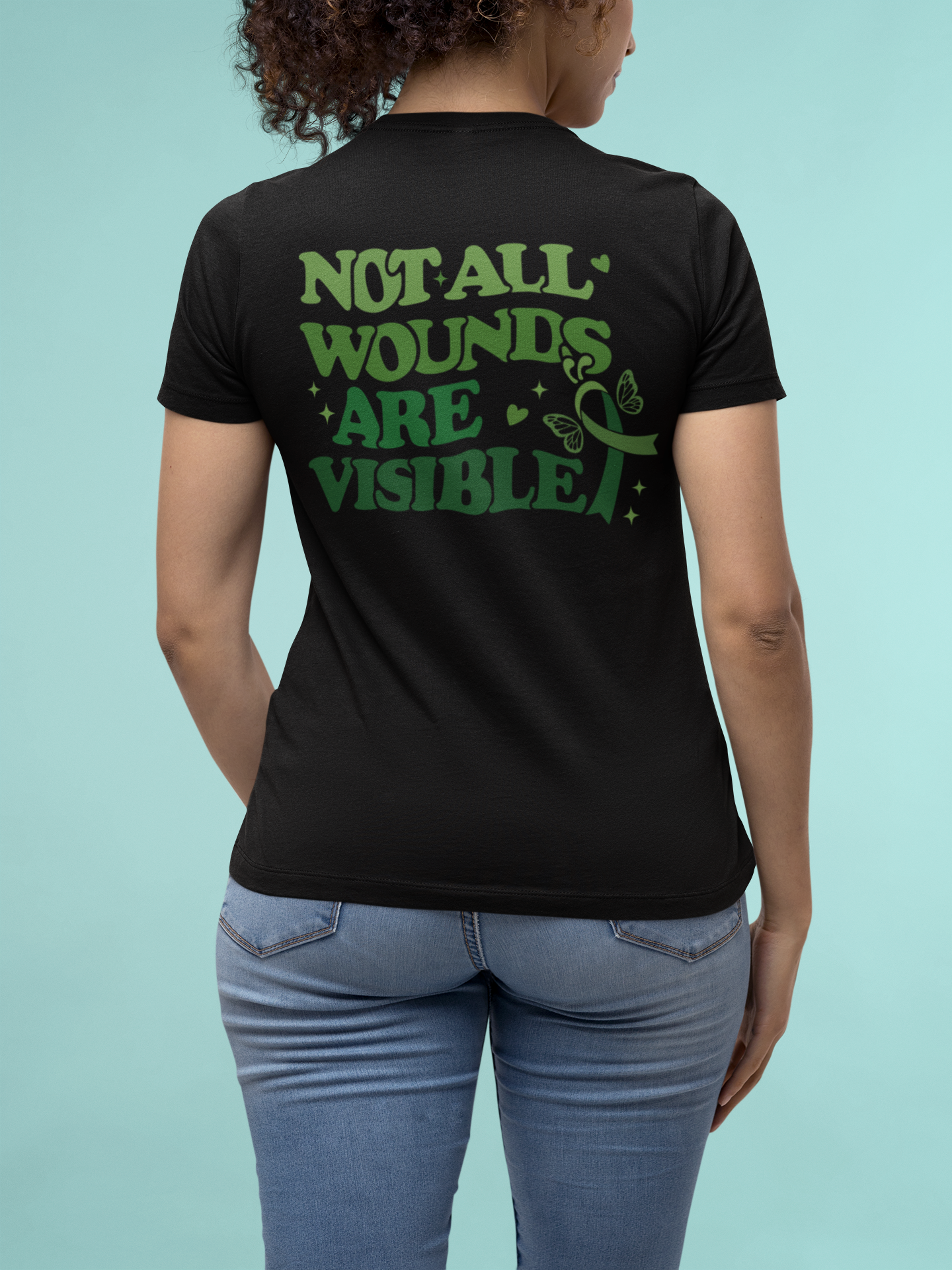 Woman wearing a black mental health awareness t-shirt with a large green “Not All Wounds Are Visible” butterfly and green ribbon design on the back.