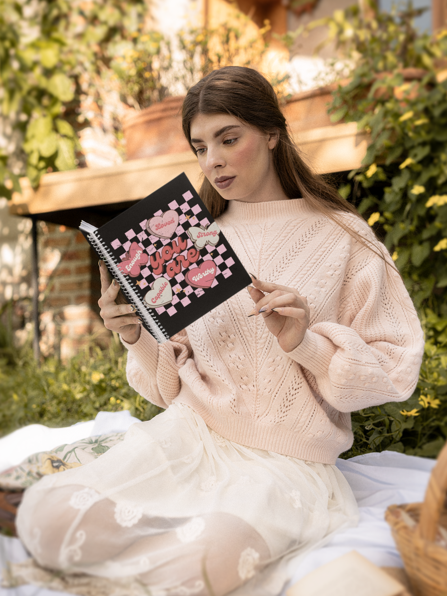 Woman in a pink sweater holding a butterfly affirmation notebook with hearts, promoting self care design outdoors