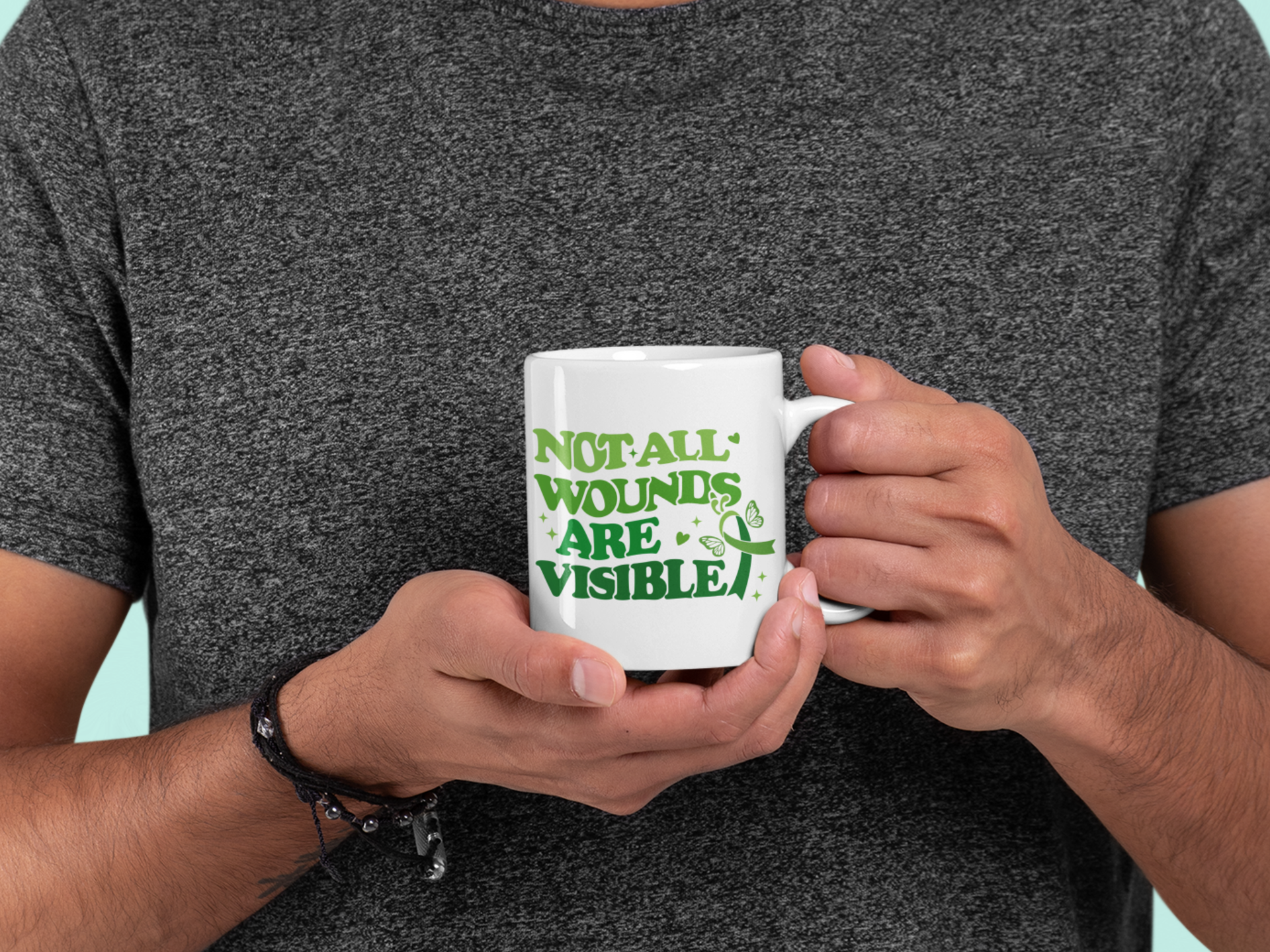 Person holding a white mental health awareness coffee mug with green “Not All Wounds Are Visible” butterfly and ribbon design.