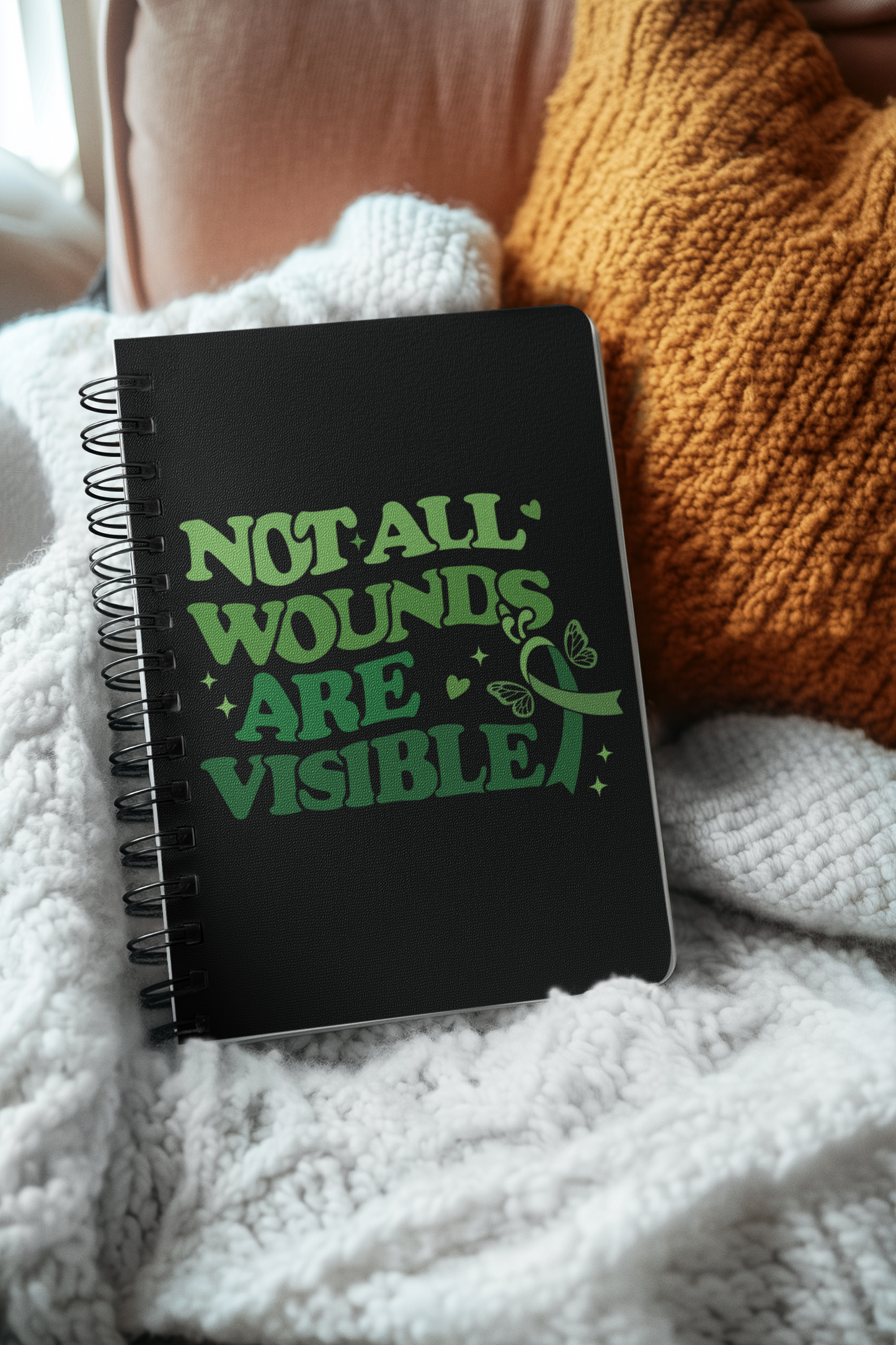 Black mental health awareness spiral notebook with green “Not All Wounds Are Visible” butterfly design placed on cozy blankets.