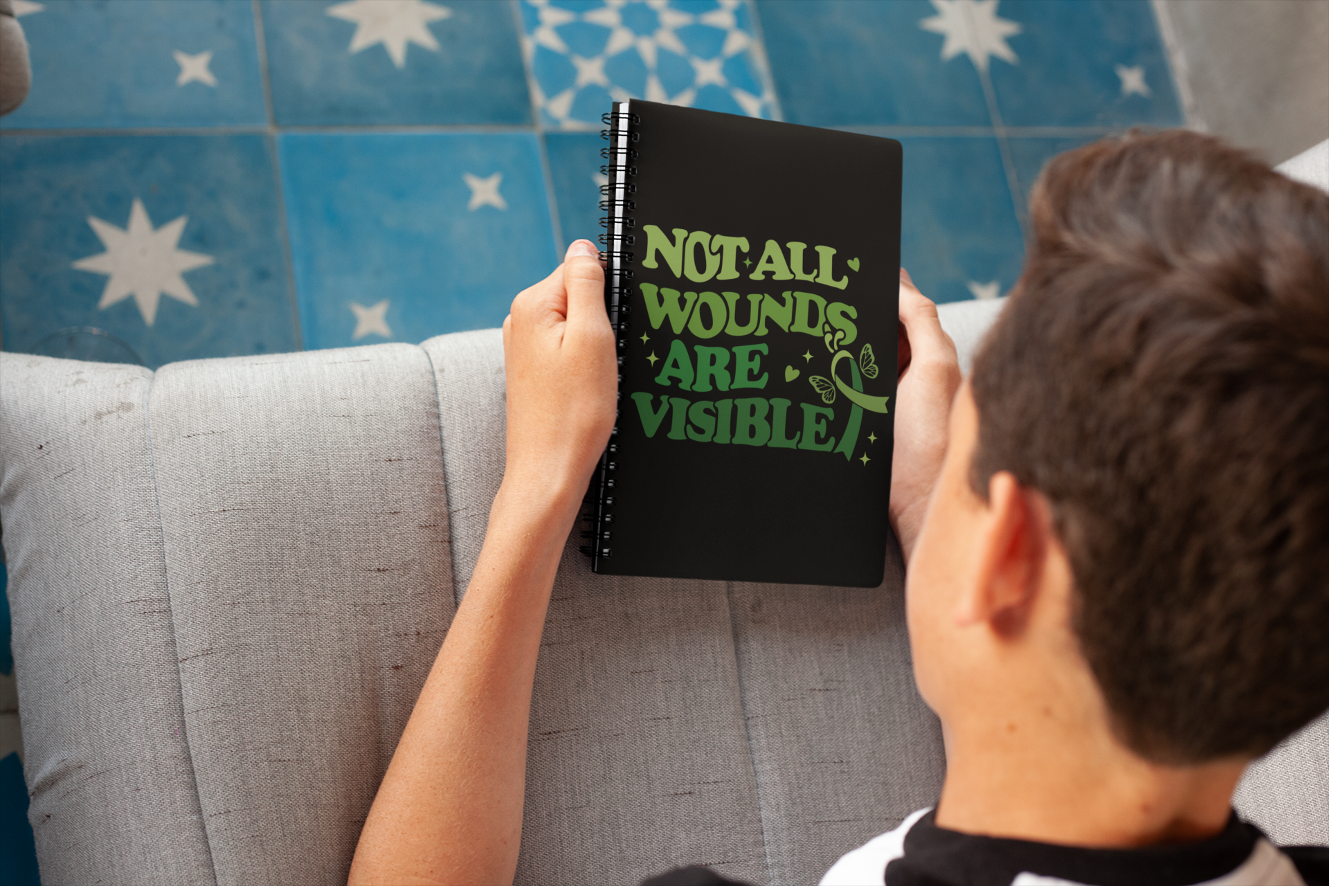 Person holding a black spiral notebook with green “Not All Wounds Are Visible” mental health awareness butterfly and ribbon design.