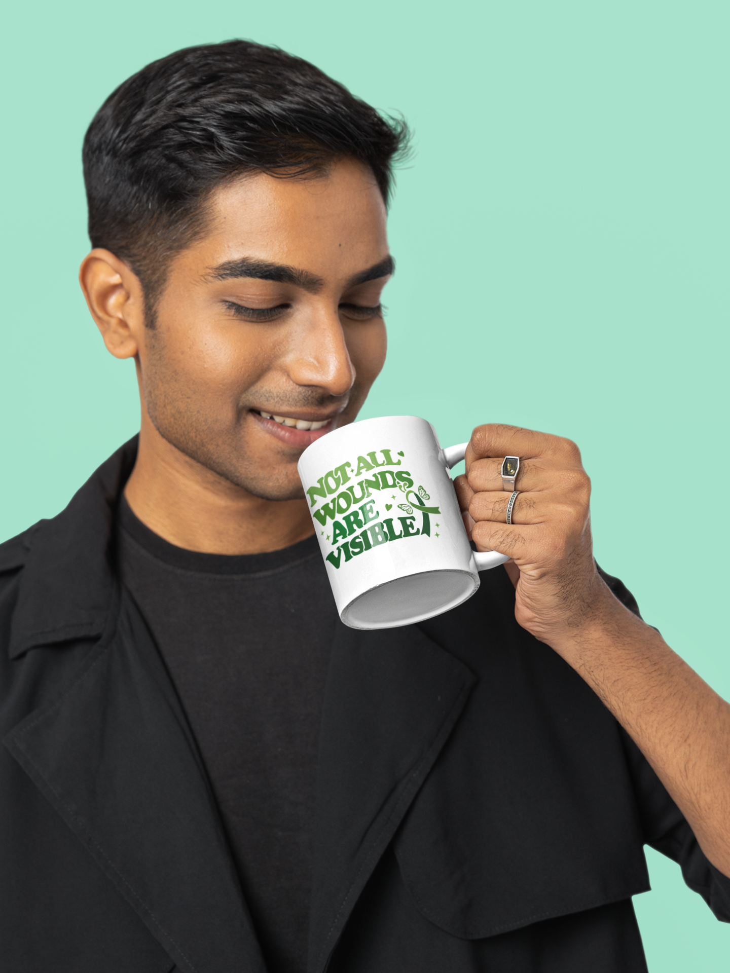 Man drinking from a white mental health awareness mug featuring a green “Not All Wounds Are Visible” butterfly and ribbon design.