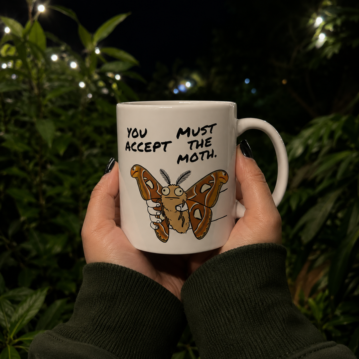 Person holding a You Must Accept The Moth Atlas Moth coffee mug outdoors at night surrounded by lush greenery and warm lights