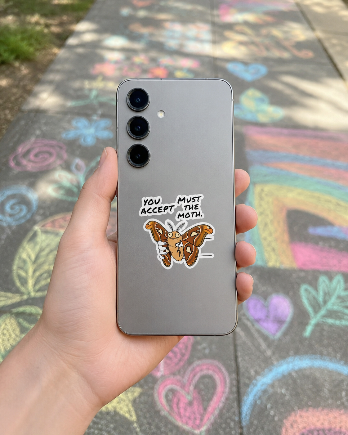 Atlas Moth vinyl sticker applied to the back of a smartphone, featuring the phrase "You Must Accept The Moth" with colorful chalk drawings in the background.