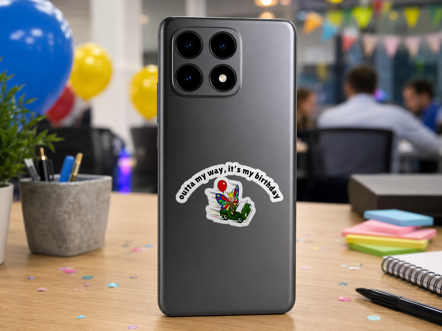 Funny birthday butterfly sticker displayed on the back of a smartphone in a festive office celebration setting.