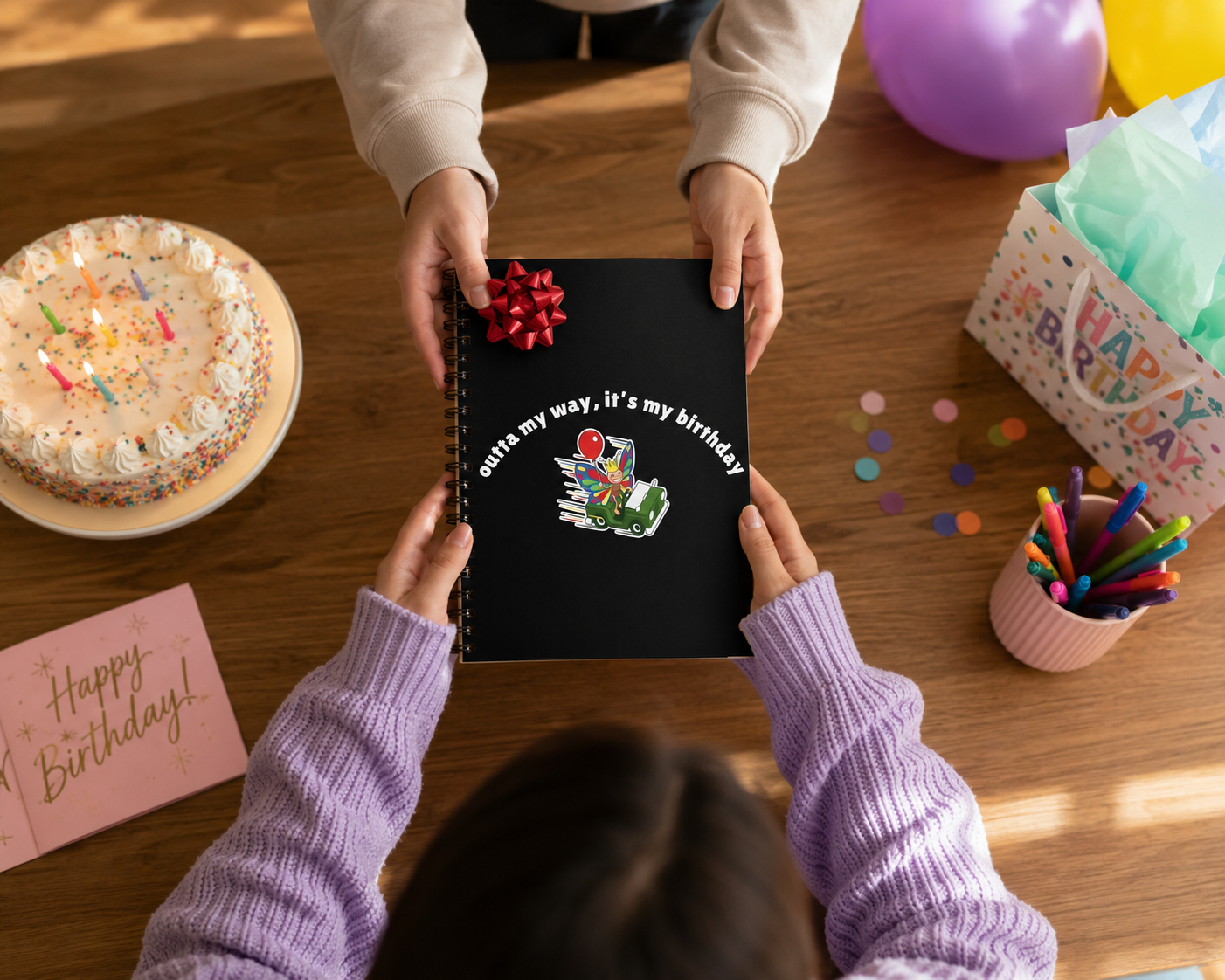 Person receiving a black butterfly birthday spiral notebook as a gift during a birthday celebration with cake, balloons, and party supplies.