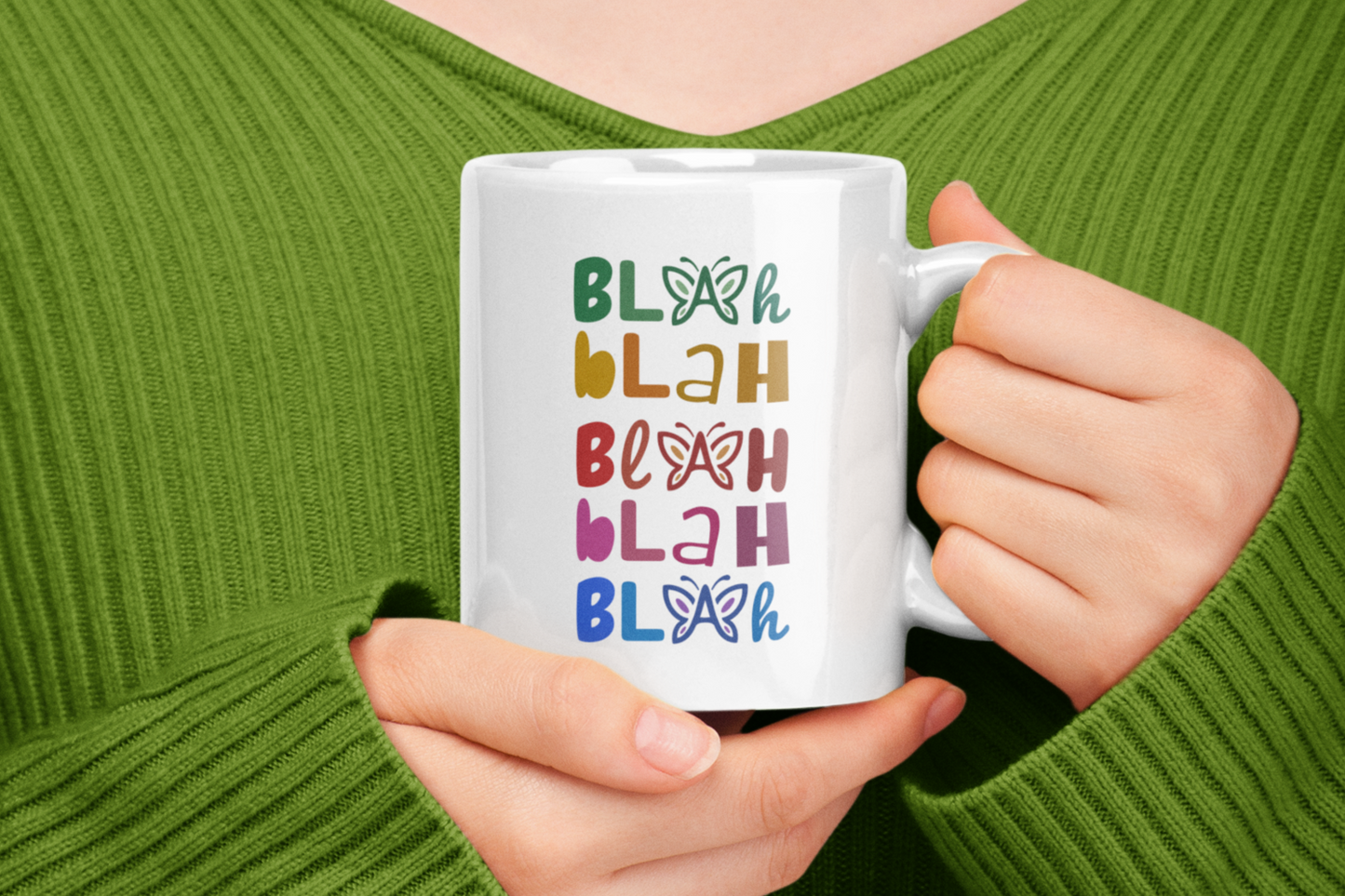 Person holding a white mug with colorful 'Blah Blah Blah' text and butterfly graphics against a green background