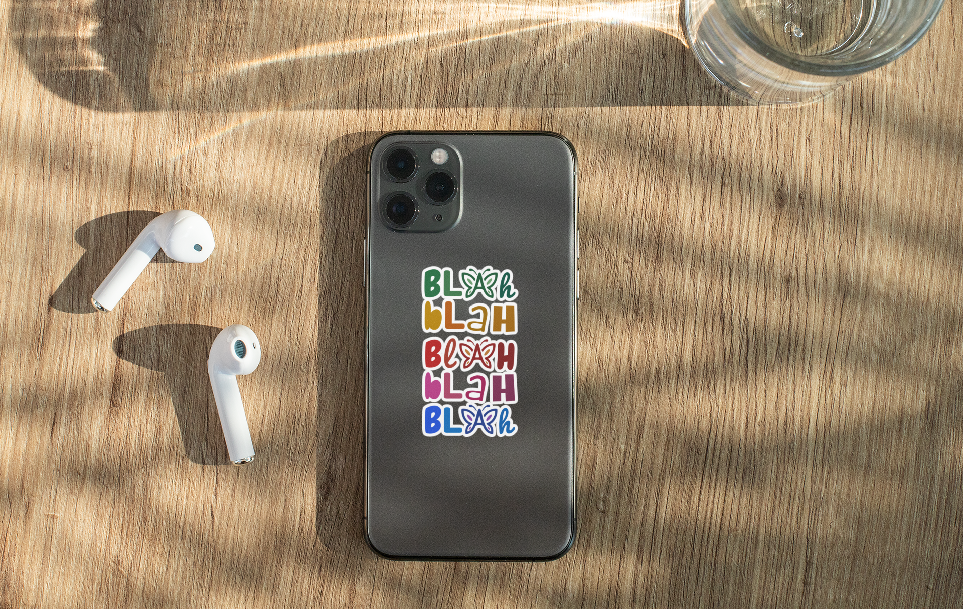 Phone with colorful 'Blah Blah Blah' text sticker on a wooden surface with airpods and a glass.