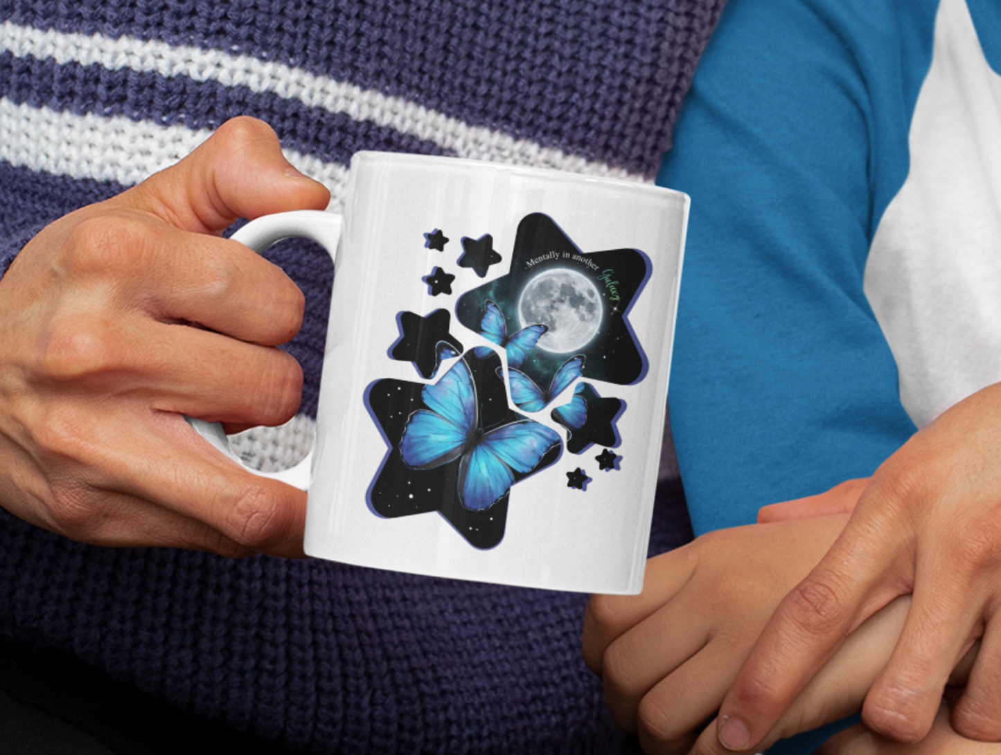 Person holding a white mug with a blue morpho butterflies with moon and stars galaxy artwork.