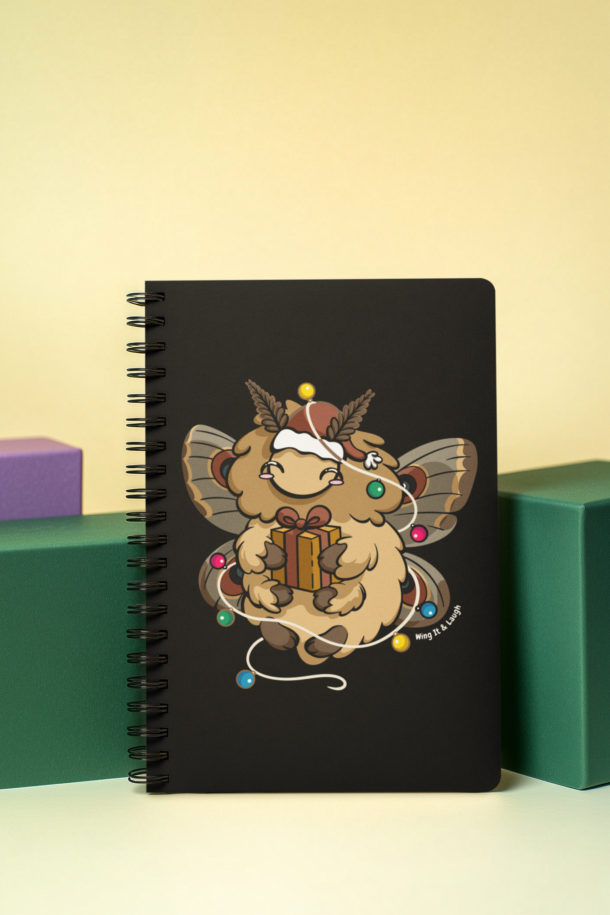 Black notebook with a cozy moth design holding a gift and surrounded by Christmas lights on a yellow background