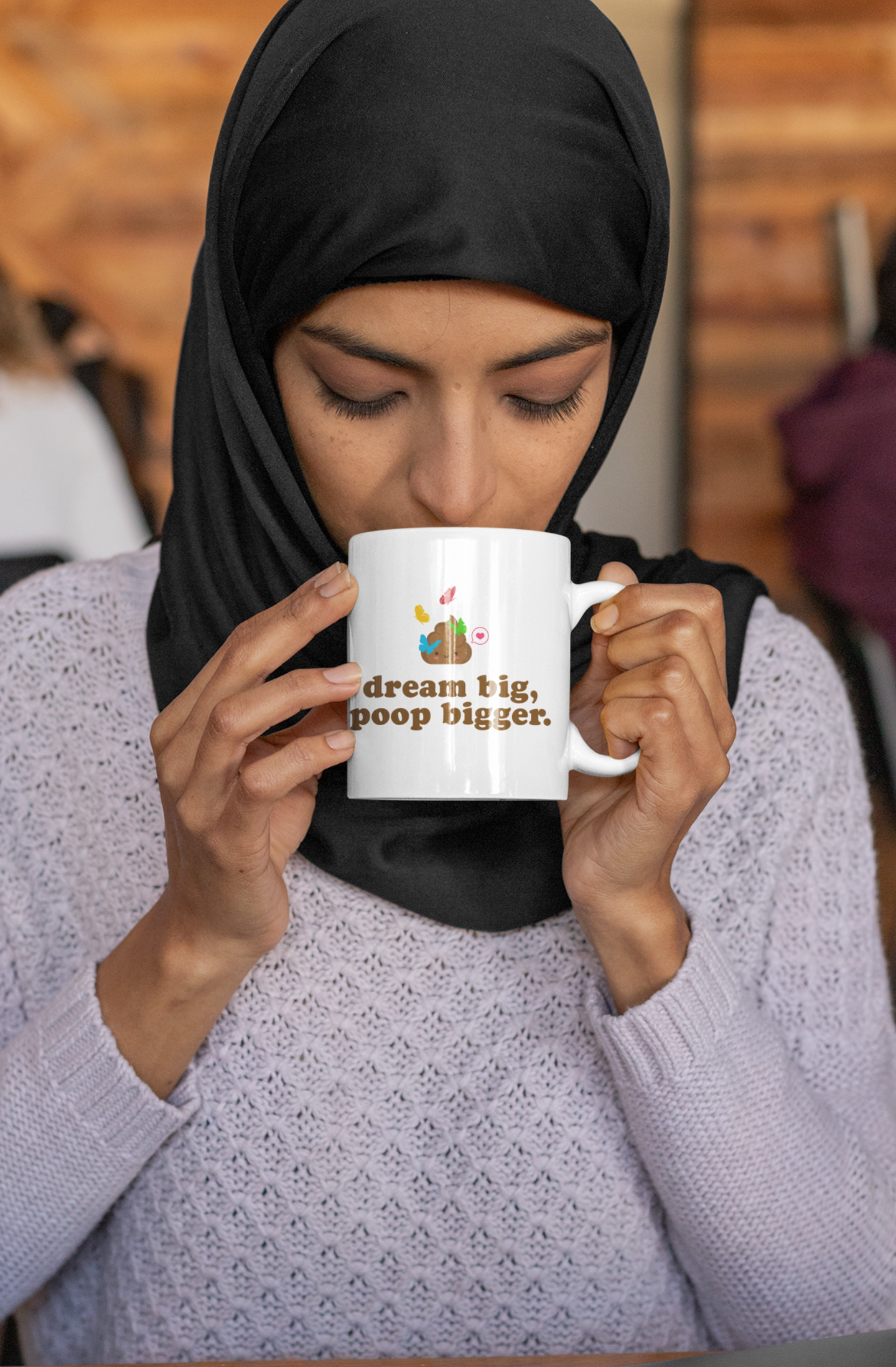 woman drinking from a "dream big, poop bigger" mug, wearing a hijab indoors