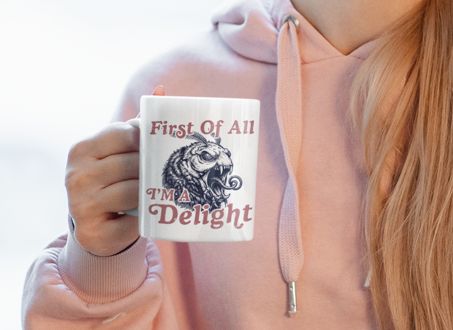 Person holding a white First Of All I’m a Delight monster moth mug while wearing a soft pink hoodie.