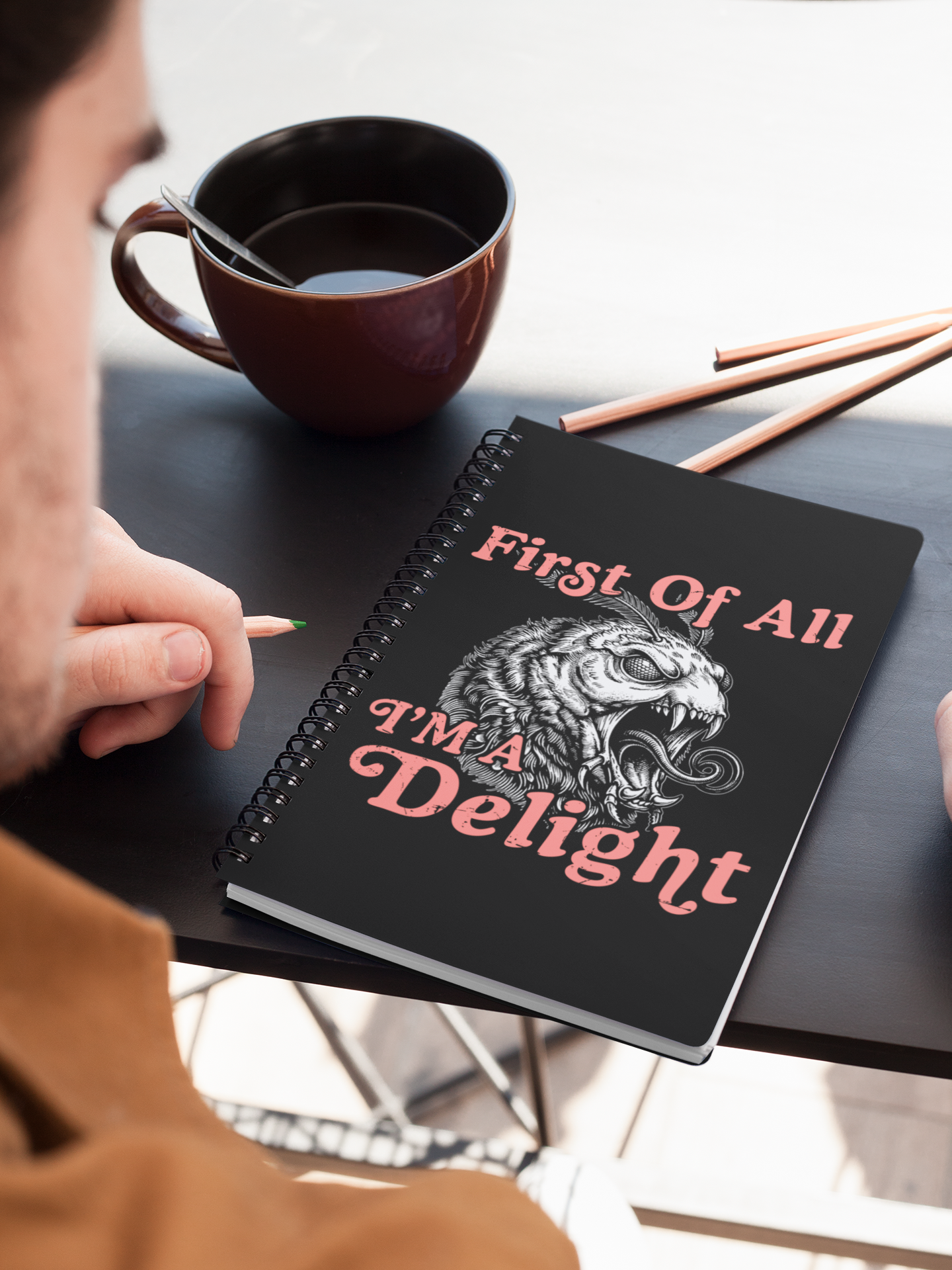 Person using a black sarcastic moth spiral notebook on a desk next to a coffee mug and pencils.