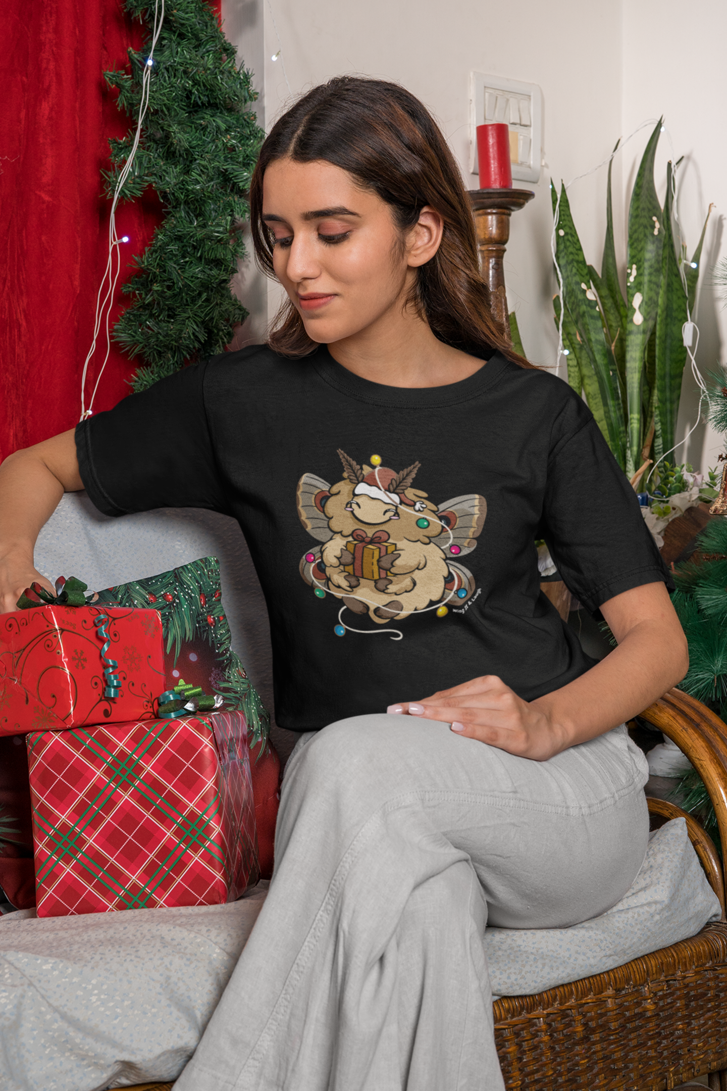 Woman sitting on a couch with Christmas decorations and wearing a black t-shirt with a cozy holiday moth holding a gift and surrounded by Christmas lights.