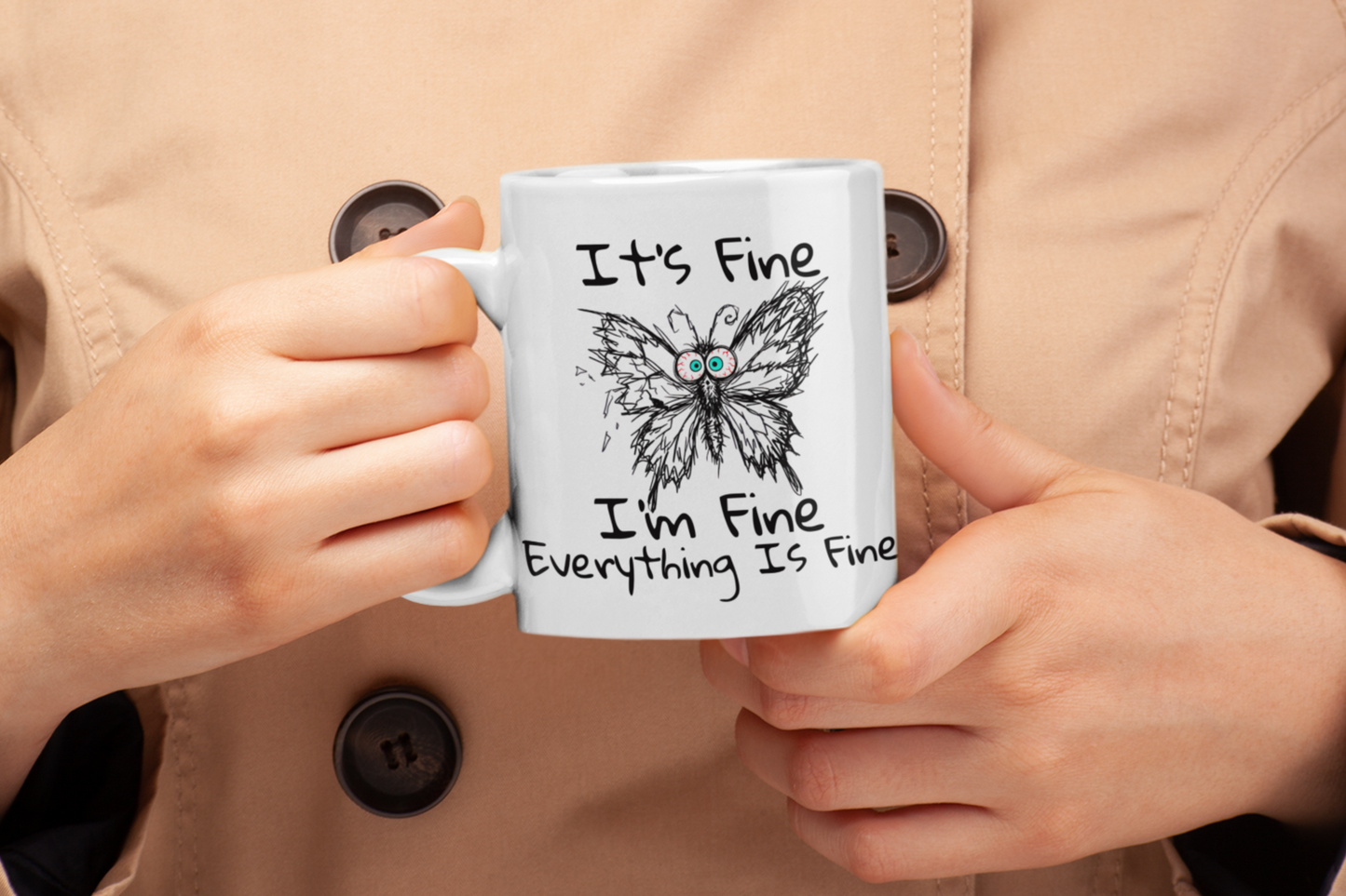 Person holding white It's Fine I'm Fine Everything Is Fine anxious butterfly coffee mug, wearing a beige coat.