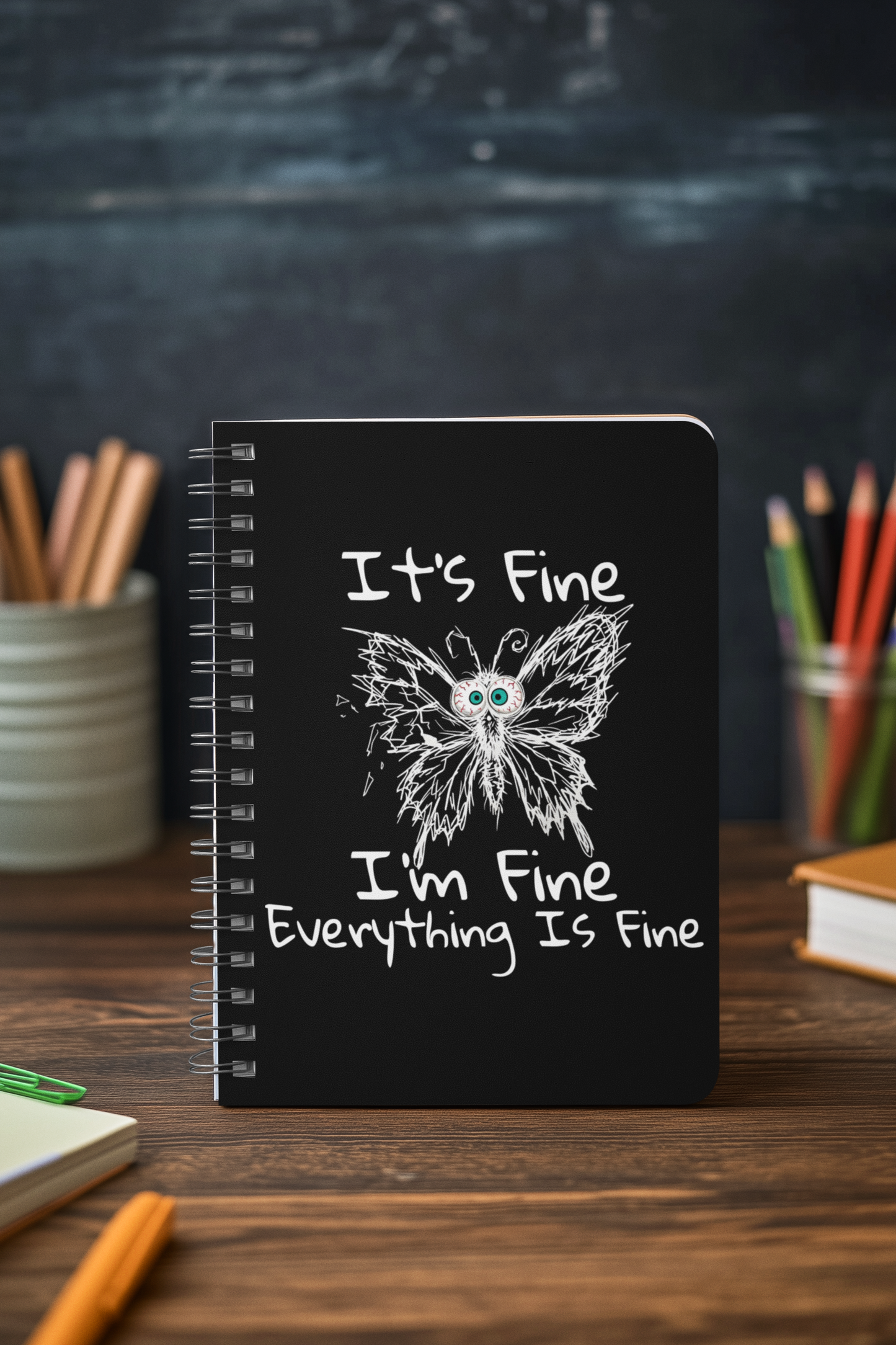 Black anxious butterfly spiral notebook standing on desk with It's Fine I'm Fine Everything Is Fine cover design with stationery items.