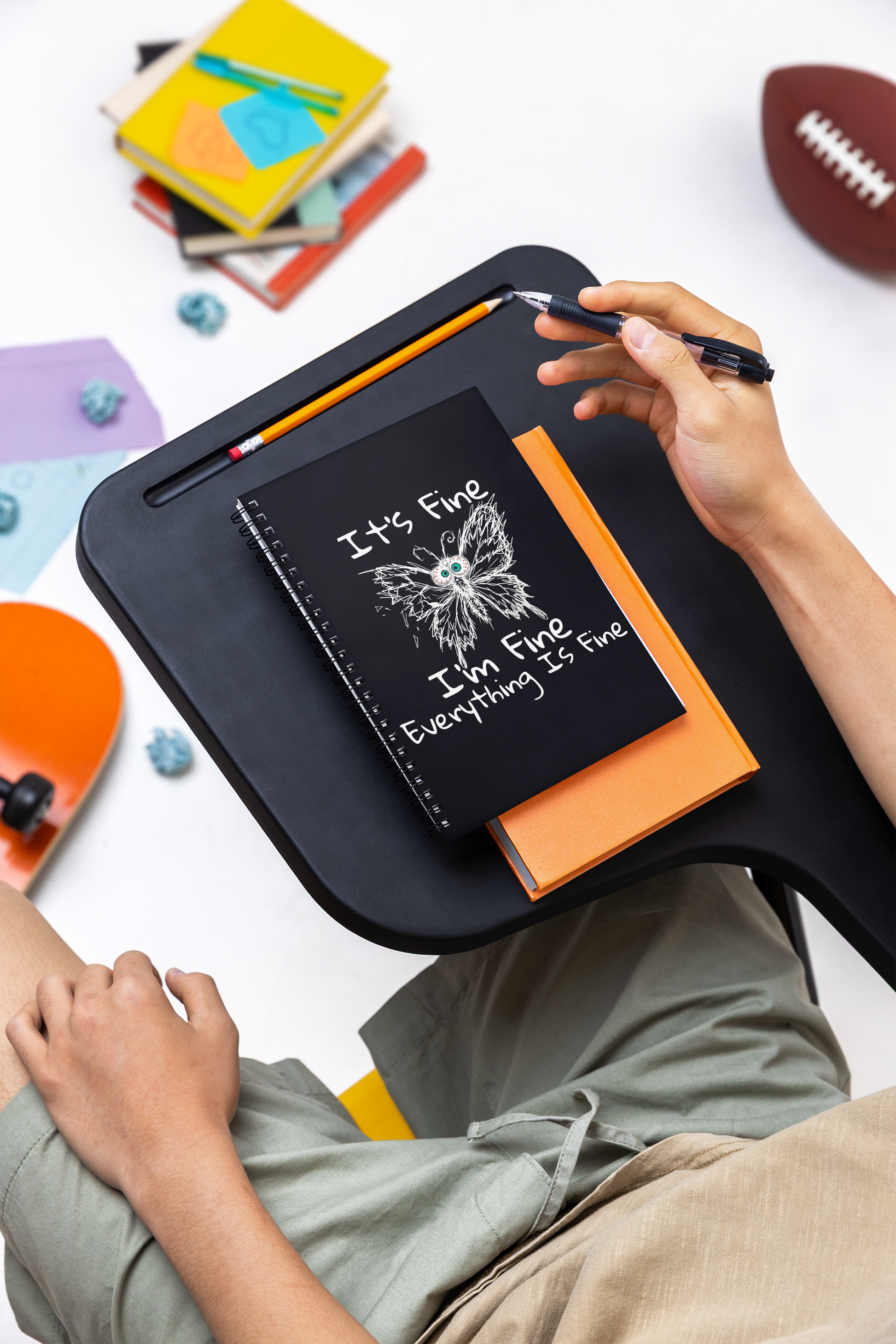 Person using It's Fine I'm Fine Everything Is Fine anxious butterfly spiral notebook on a desk, surrounded by colorful items on a white surface.