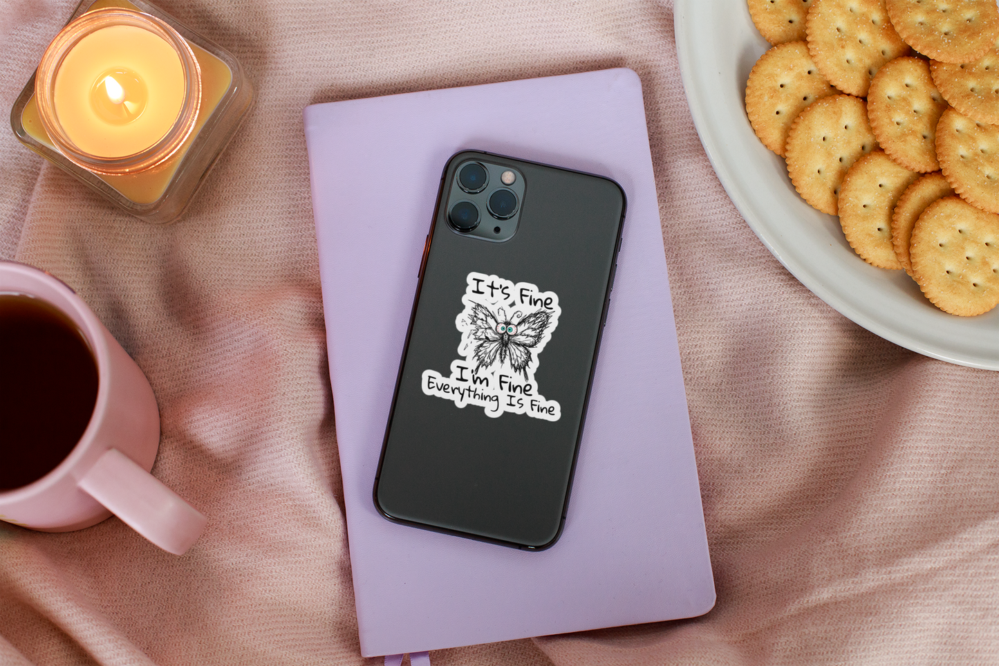 It's Fine I'm Fine Everything Is Fine anxious butterfly sticker displayed on phone case on a pink surface with a candle and cookies.