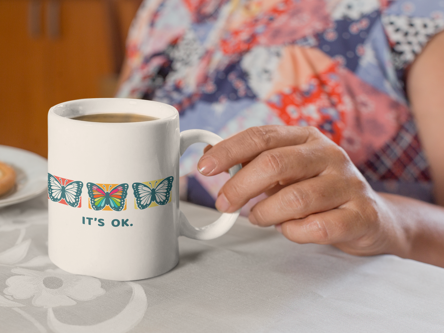 person holding a mug with butterfly design and 'it's ok' text on a table during a coffee break
