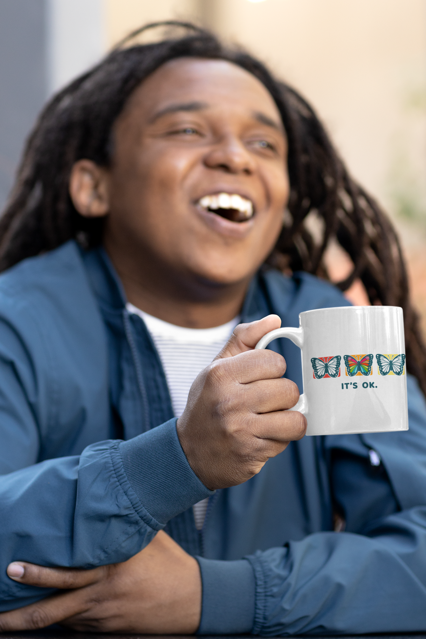 Man holding a mug with colorful butterfly design and 'it’s ok' text, laughing outdoors.