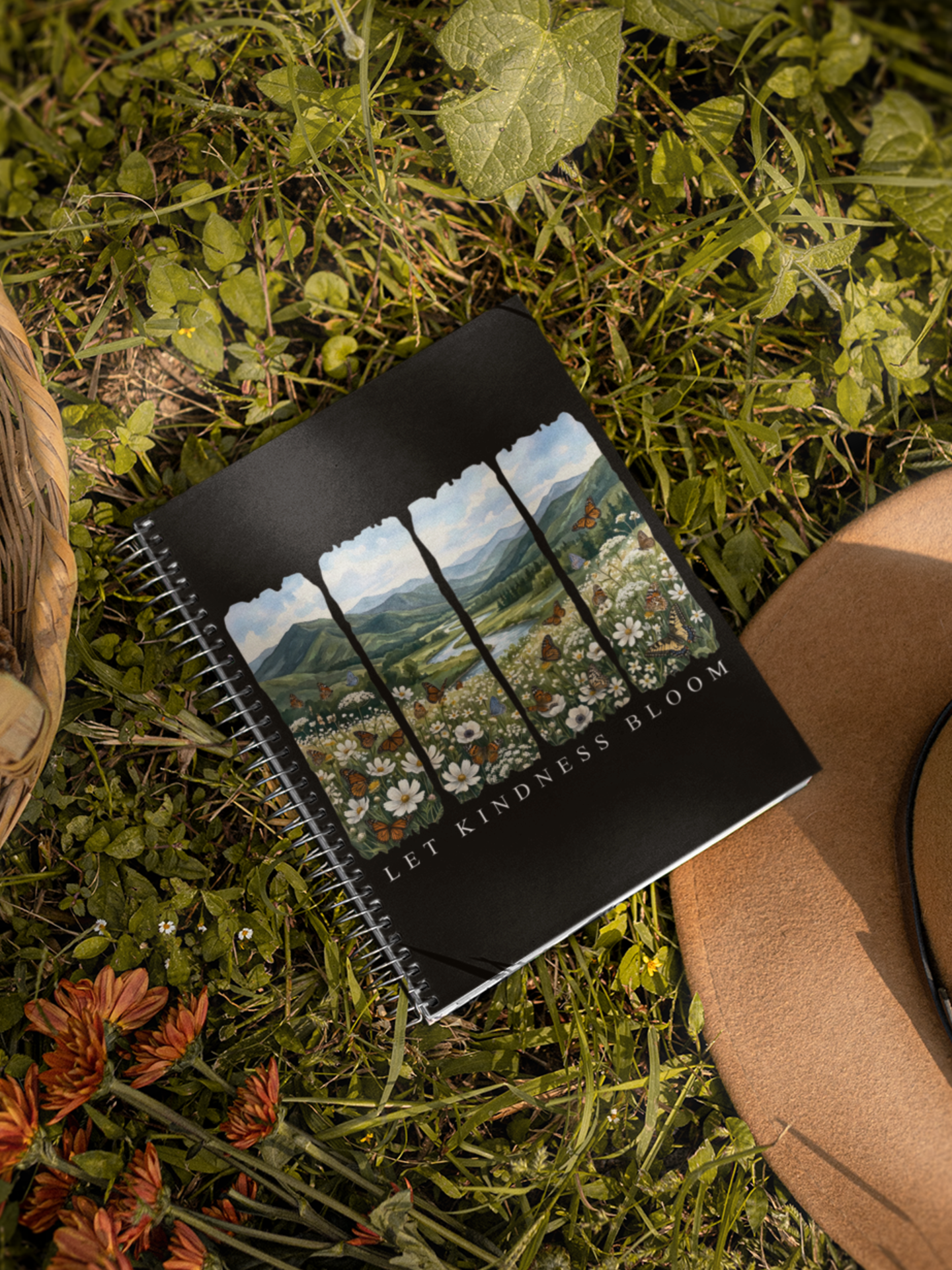 Let Kindness Bloom butterfly floral spiral notebook shown outdoors on grass with a hat and flowers