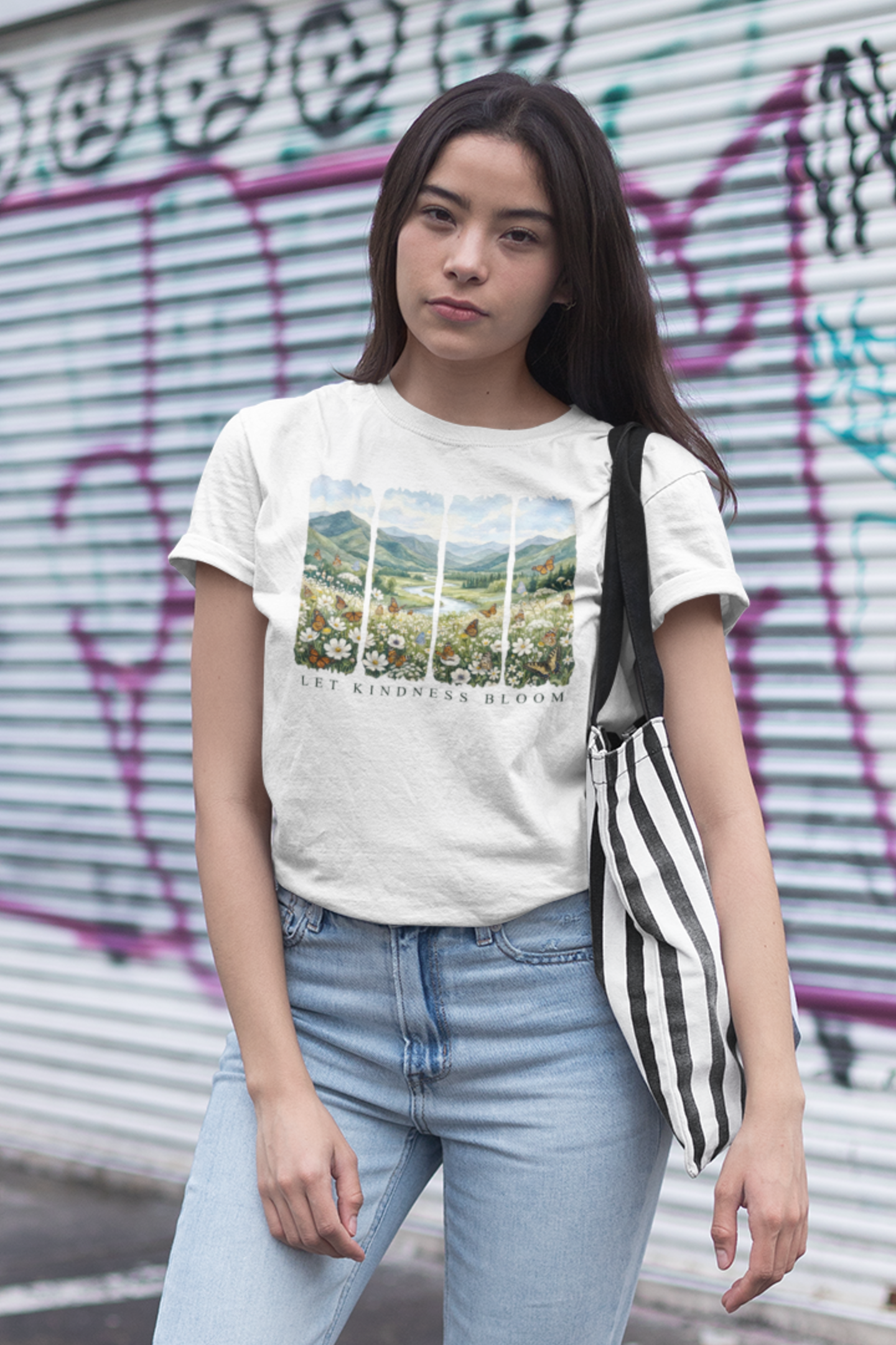 Woman wearing a white Let Kindness Bloom butterfly and floral landscape t-shirt in front of a graffiti-covered wall.