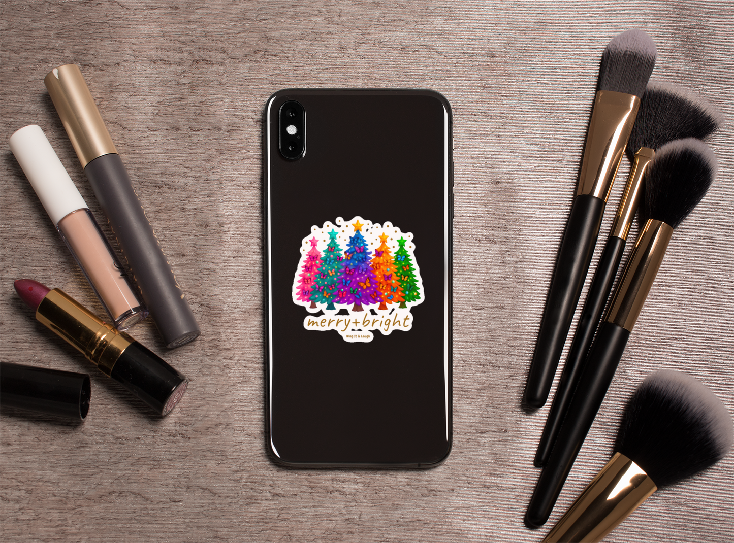 Merry and bright Christmas trees butterfly sticker on phone case on a wooden surface with makeup items.