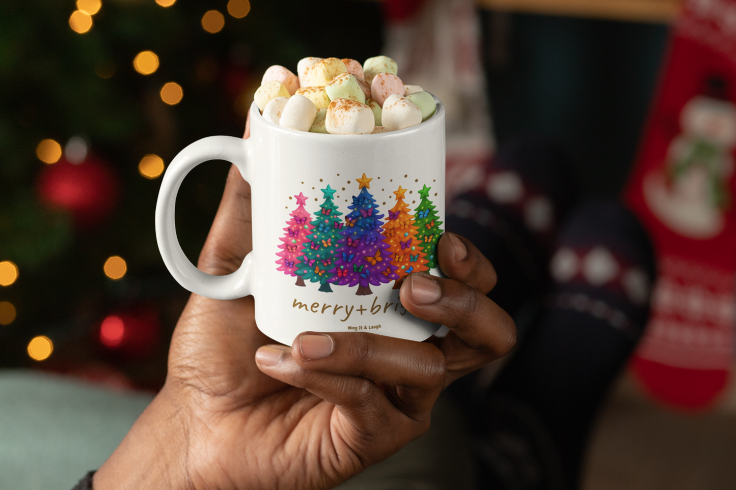 Hand holding a mug with Christmas trees design and 'merry + bright' text, filled with cocoa and marshmallows