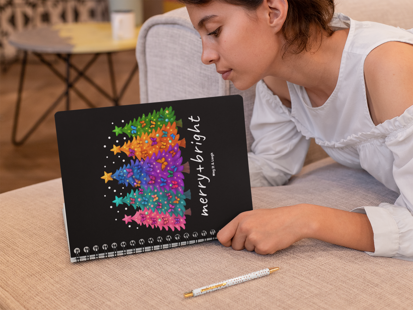 Person holding a 6x8 journal with a colorful Christmas trees design and 'merry + bright' text, on a couch.