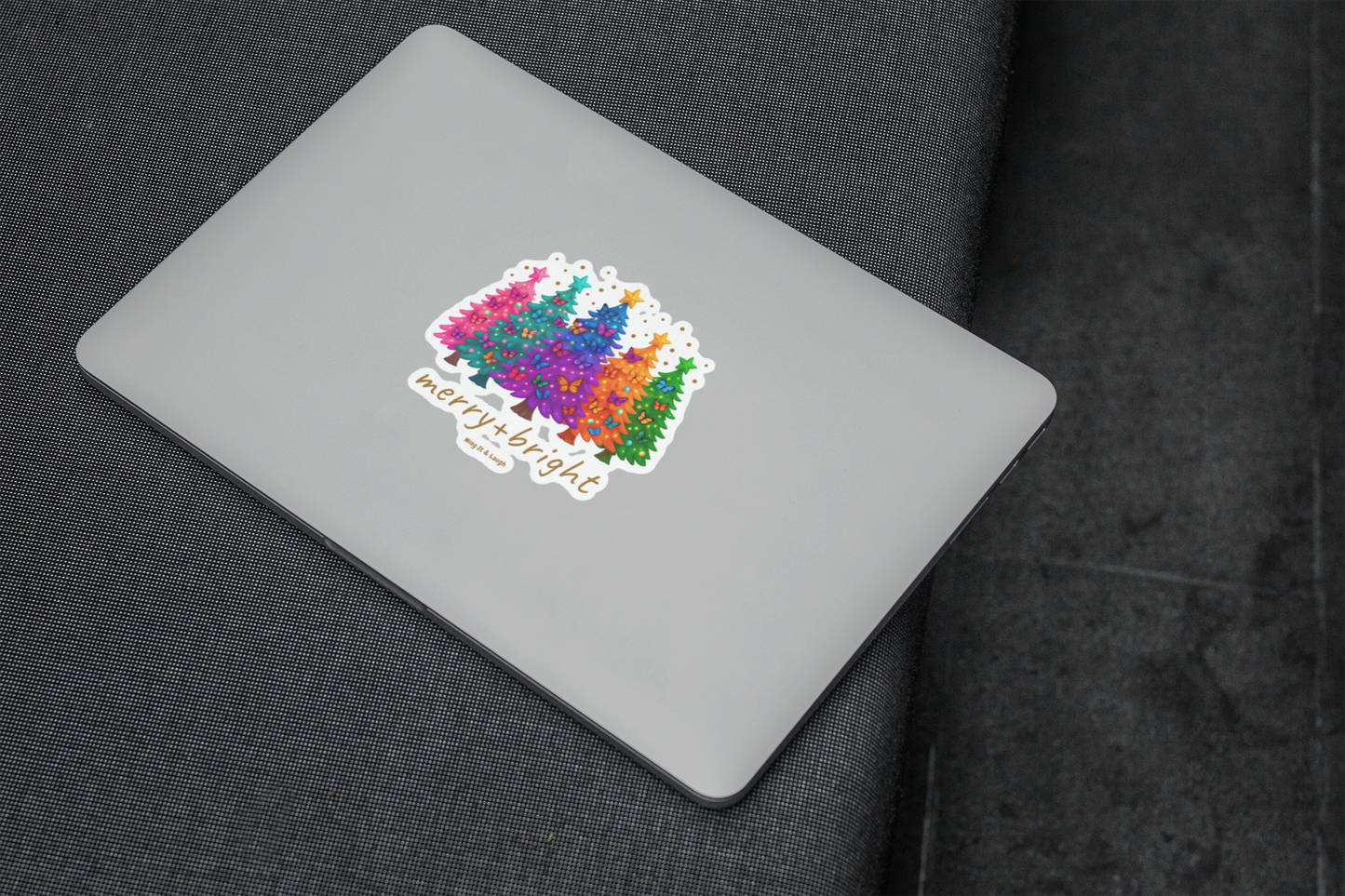 Colorful Christmas trees and butterflies sticker with text 'merry & bright' on a laptop on a dark fabric background