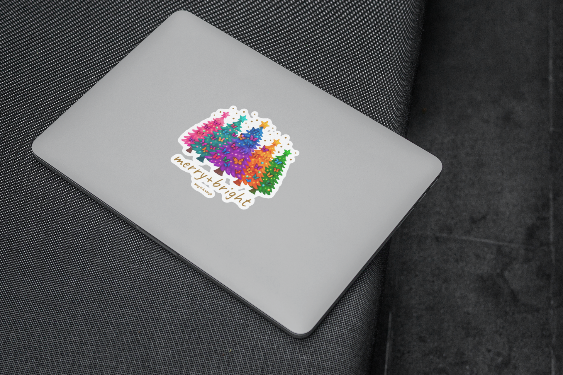 Colorful Christmas trees and butterflies sticker with text 'merry & bright' on a laptop on a dark fabric background