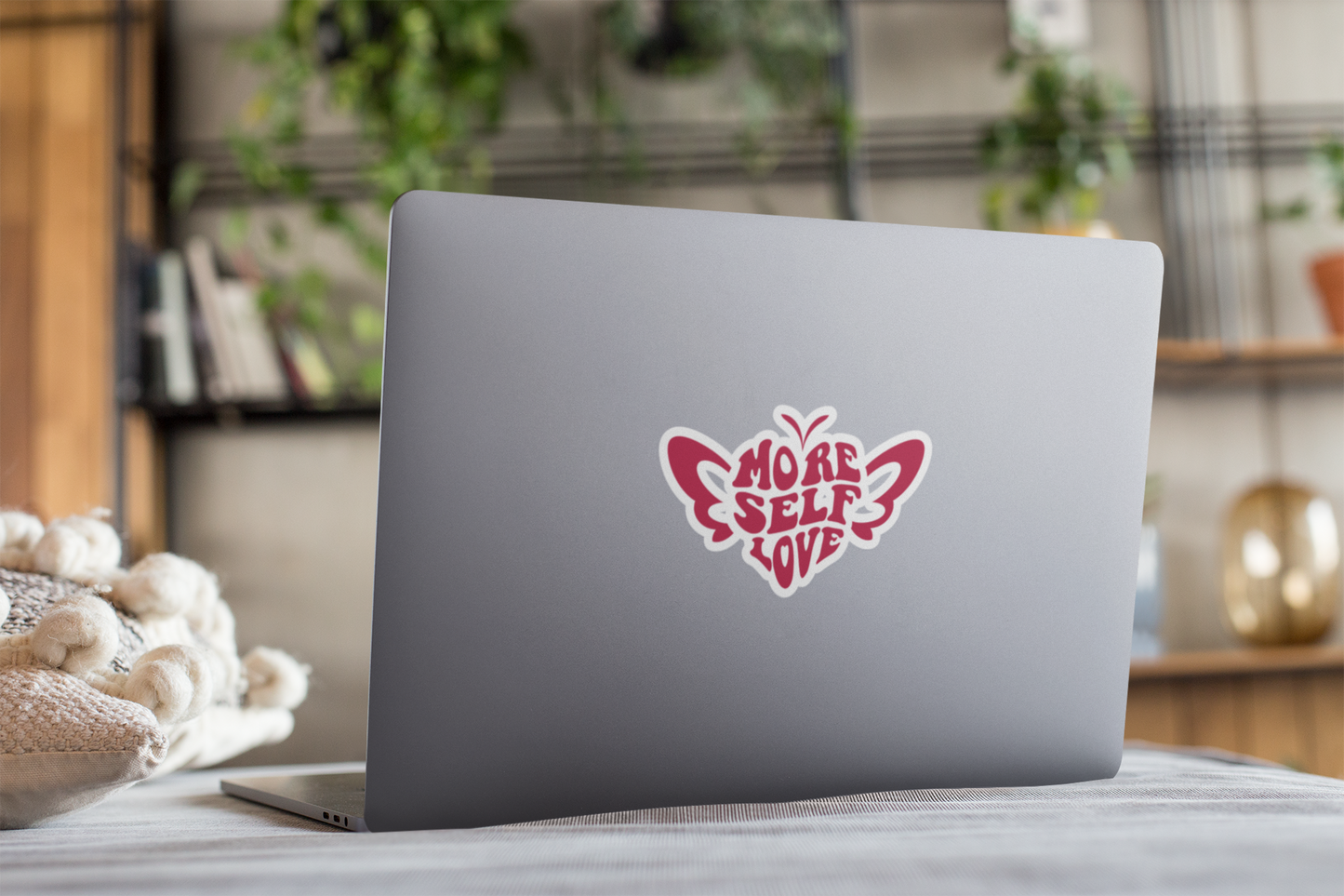 Laptop with a More Self Love self care butterfly sticker on a surface with a blurred indoor background