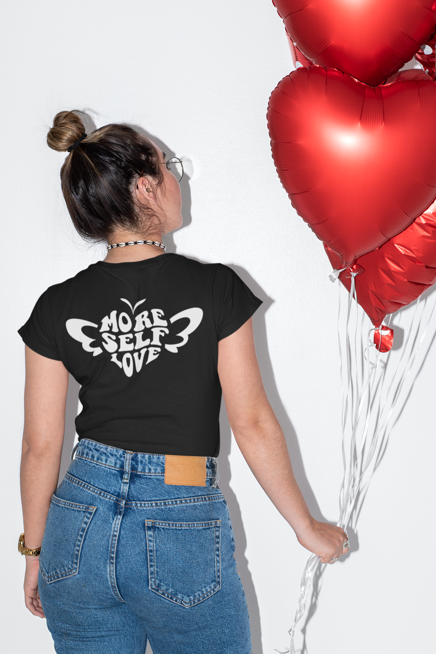 Person wearing black More Self Love butterfly Valentine shirt, holding red heart-shaped balloons.