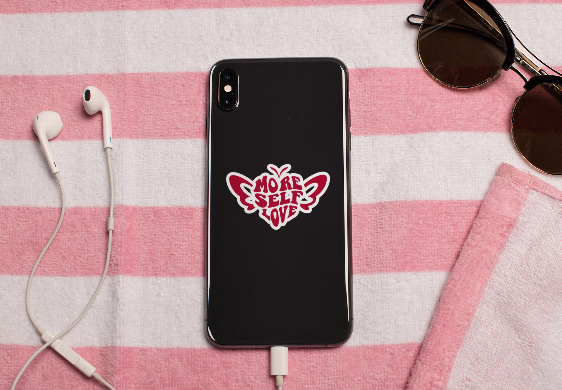 Black phone with More Self Love butterfly sticker on a pink and white checkered background with earphones and sunglasses.