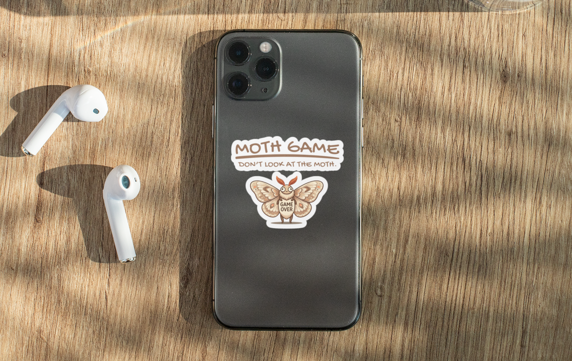 Funny Moth Game sticker applied to a phone, showing the cute moth Game Over design and slogan on a wooden surface with white earbuds nearby