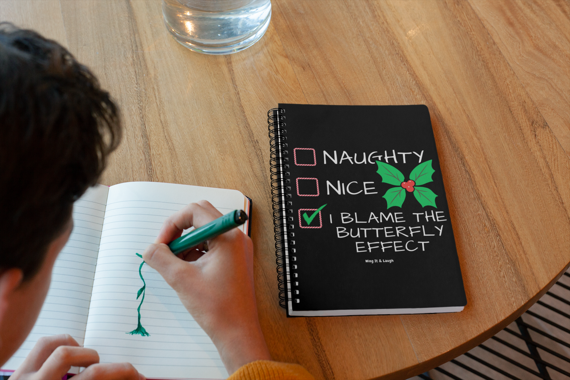 Person writing in a naughty or nice butterfly effect spiral notebook at a wooden table.
