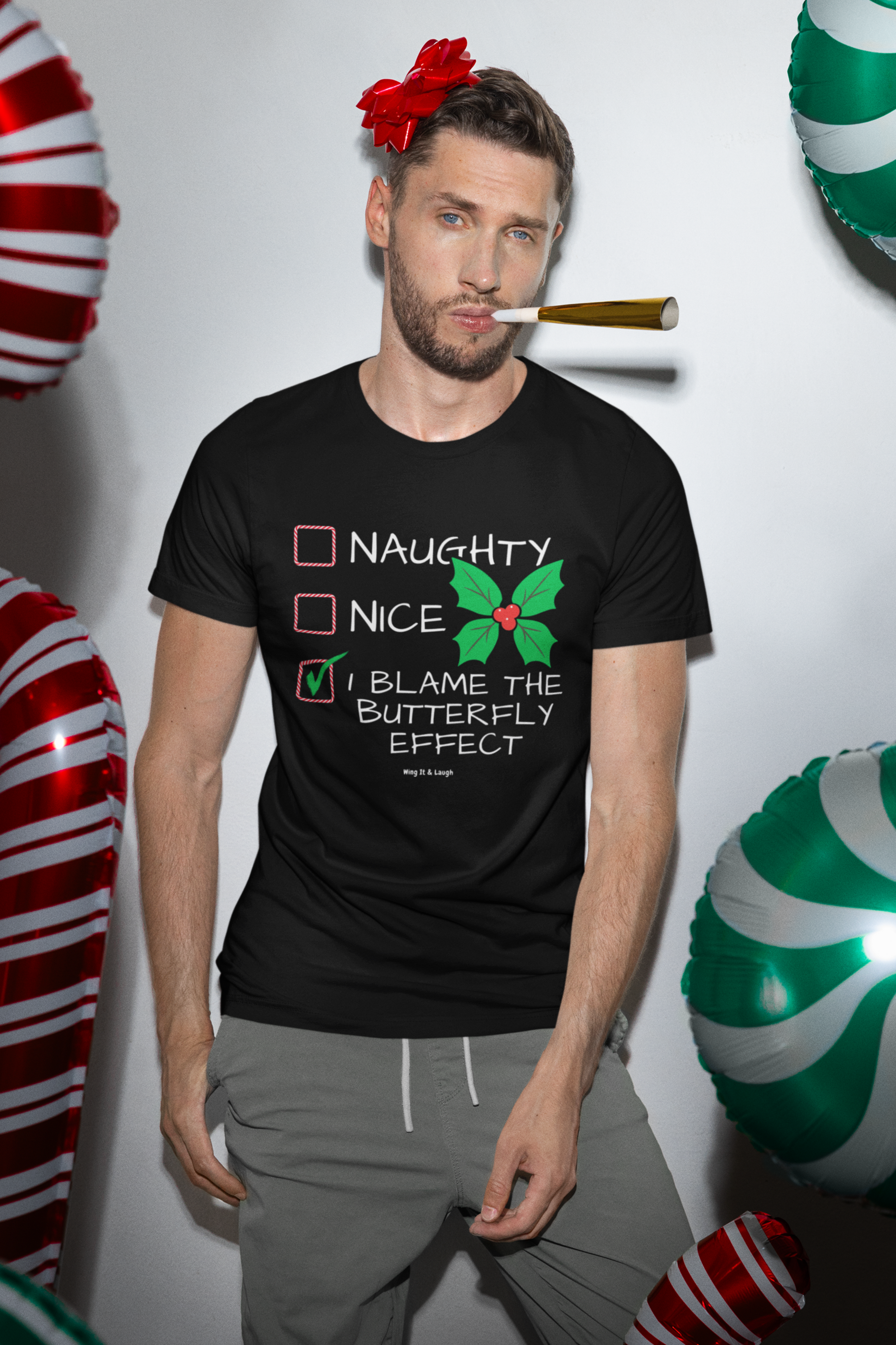 Man wearing a black “I Blame the Butterfly Effect” naughty or nice Christmas checklist t-shirt, standing against a white wall with festive balloons.