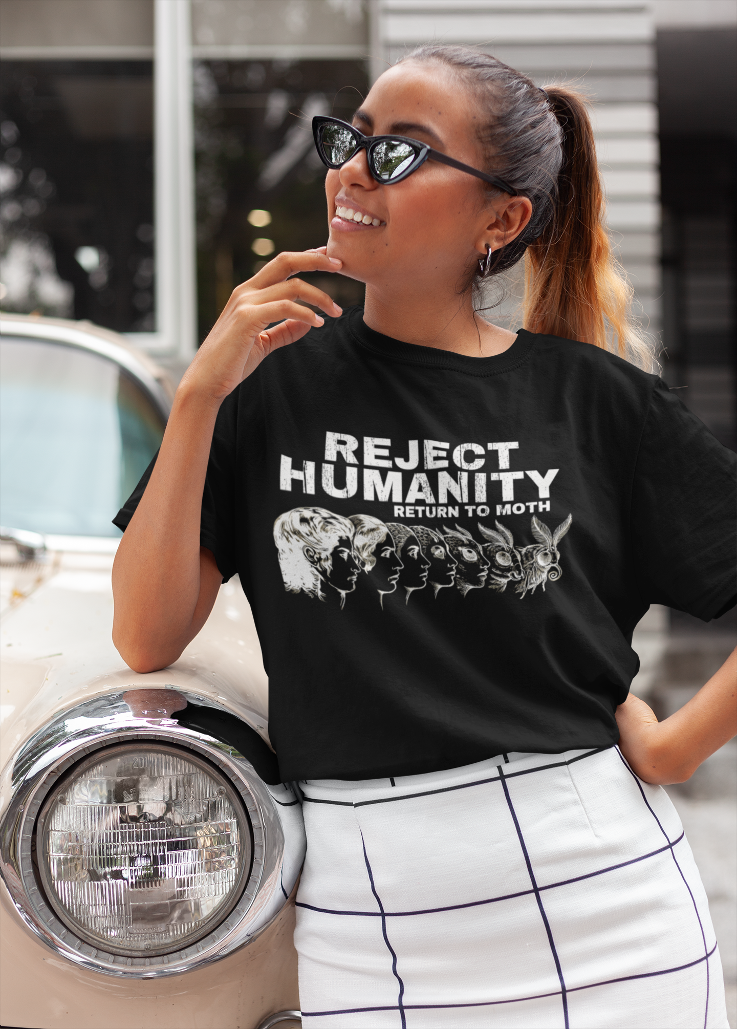Woman wearing funny moth meme shirt “Reject Humanity Return to Moth”, standing next to a vintage car.