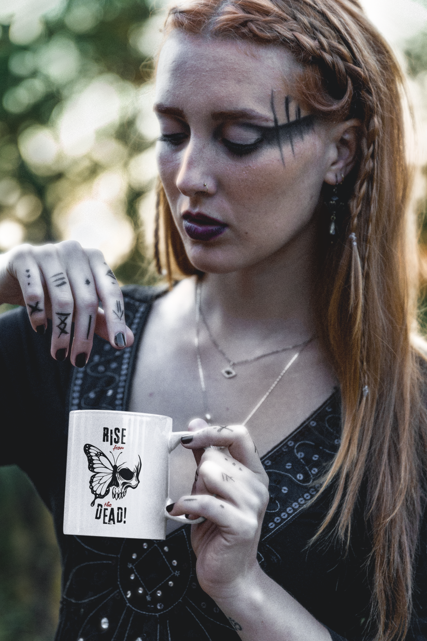 Goth woman with tattoos holding a mug with a “Rise from the Dead!” text and a skull-butterfly design.