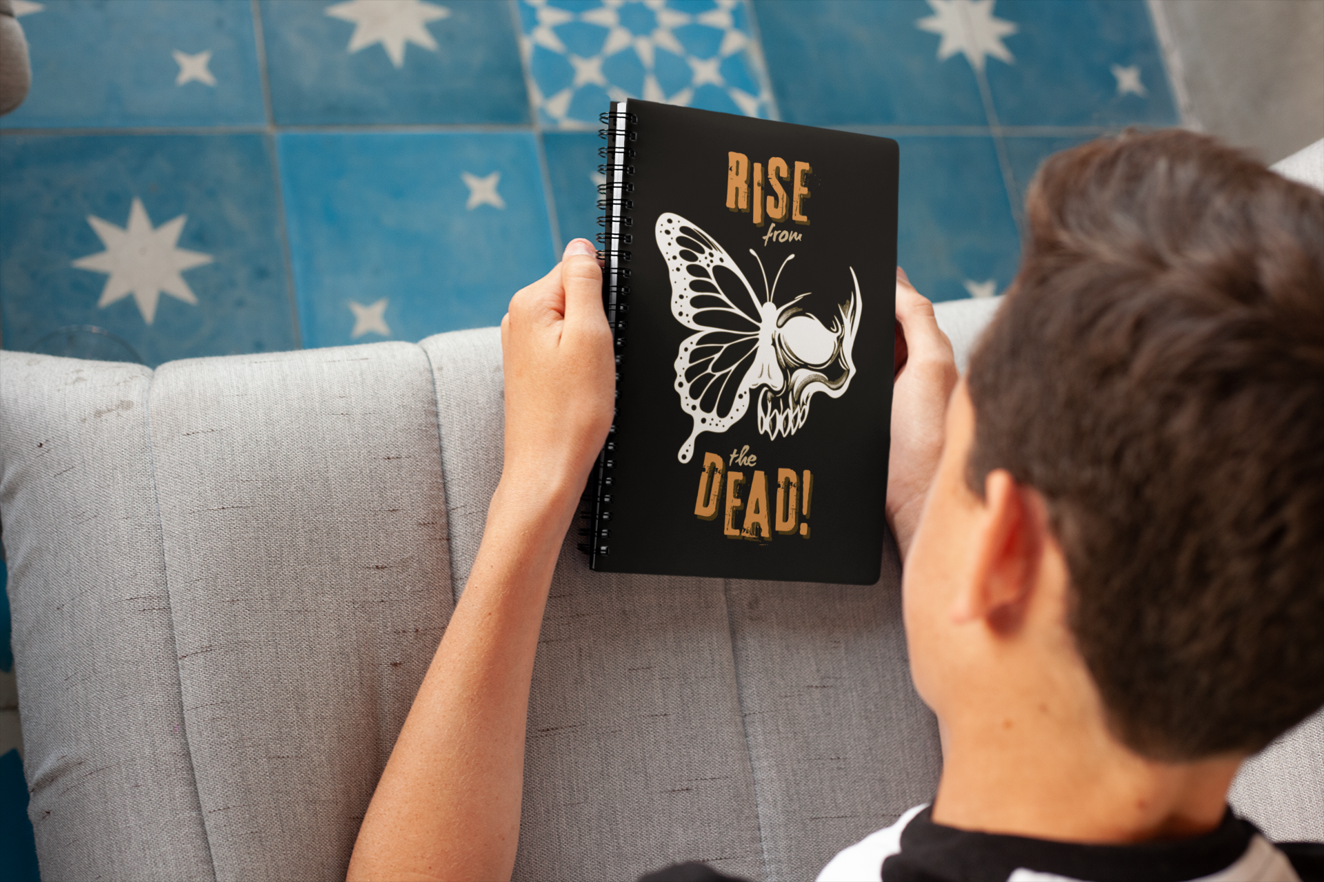 Person holding a skull-butterfly 6×8 journal, sitting on a couch against a blue patterned wall.