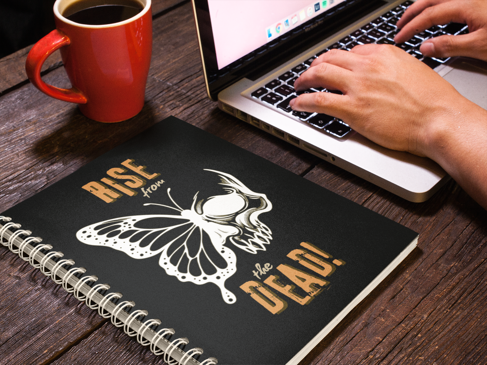 Person using a laptop with a skull-butterfly spiral notebook on a wooden desk.