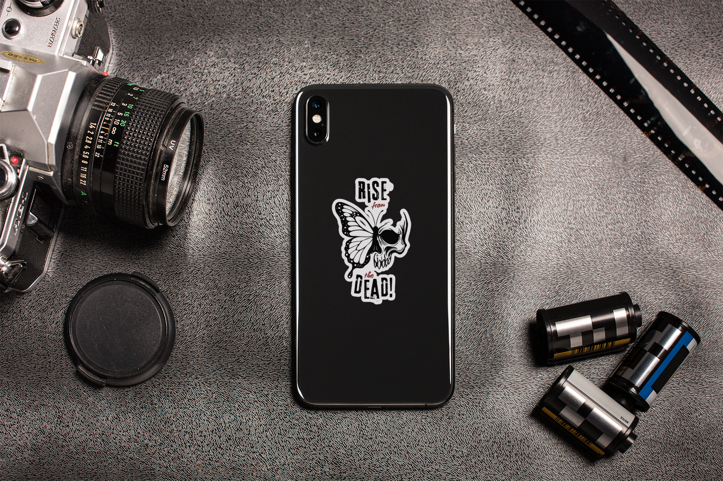 Skull-butterfly sticker on a black phone, lifestyle shot with camera gear