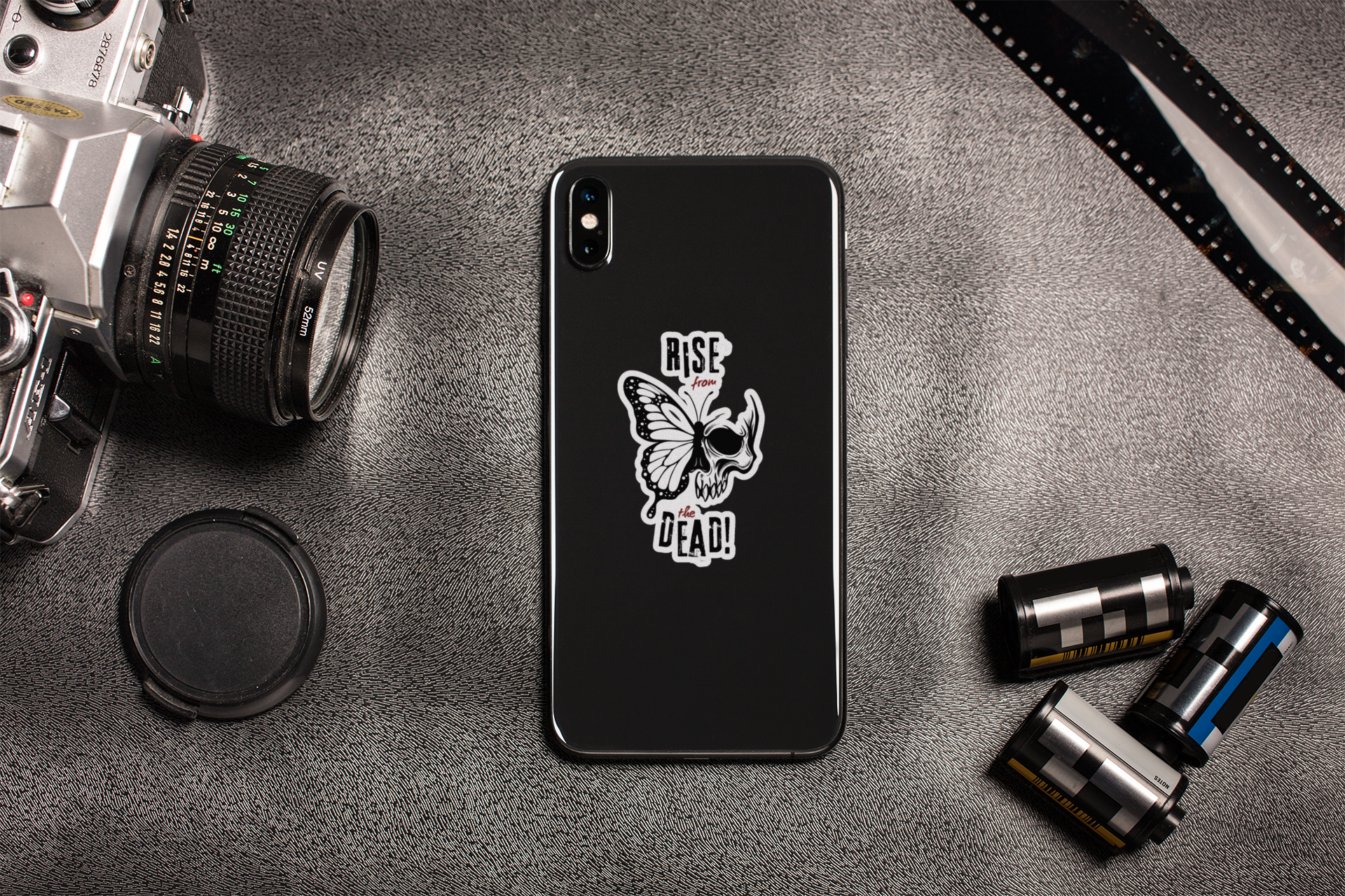 Skull-butterfly sticker on a black phone, lifestyle shot with camera gear