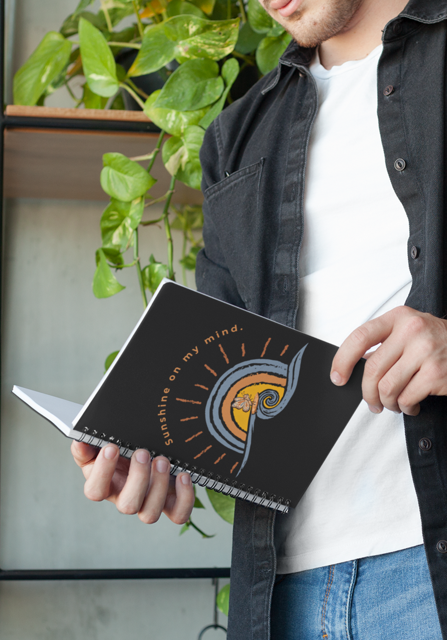 Person holding Sunshine On My Mind butterfly notebook with black cover and retro butterfly wave design, surrounded by green plants.