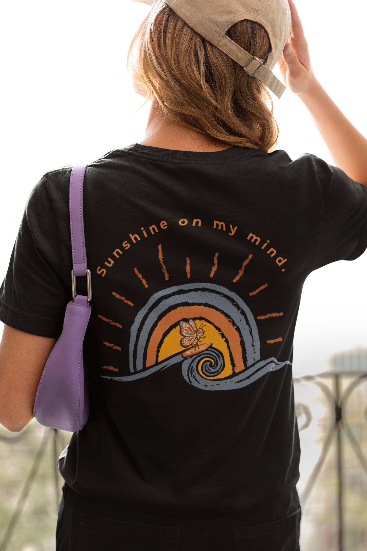 Woman wearing black Sunshine On My Mind butterfly shirt showing large retro back graphic, holding a purple bag.