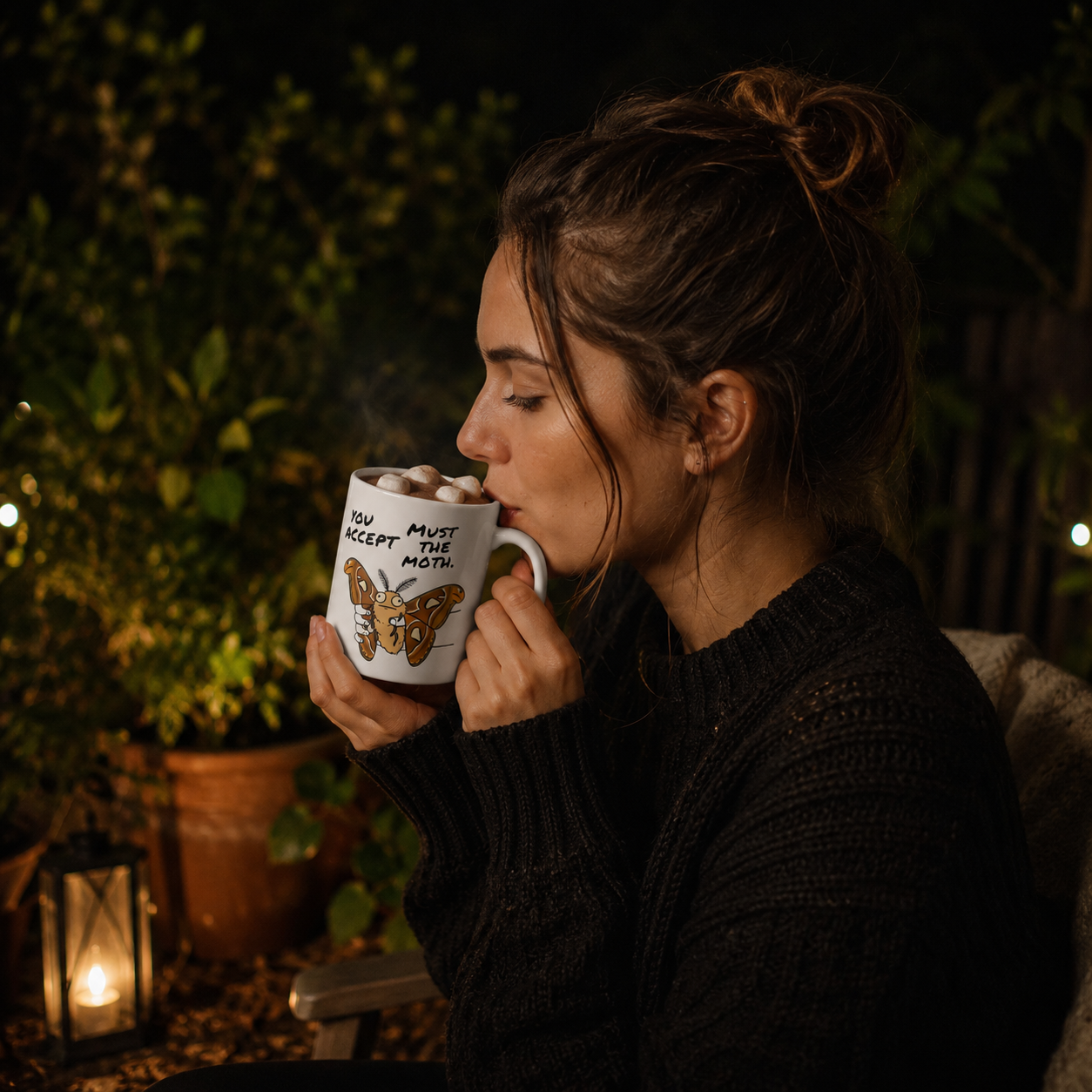 Woman drinking from a funny Atlas Moth mug featuring the phrase You Must Accept The Moth in a cozy nighttime garden setting