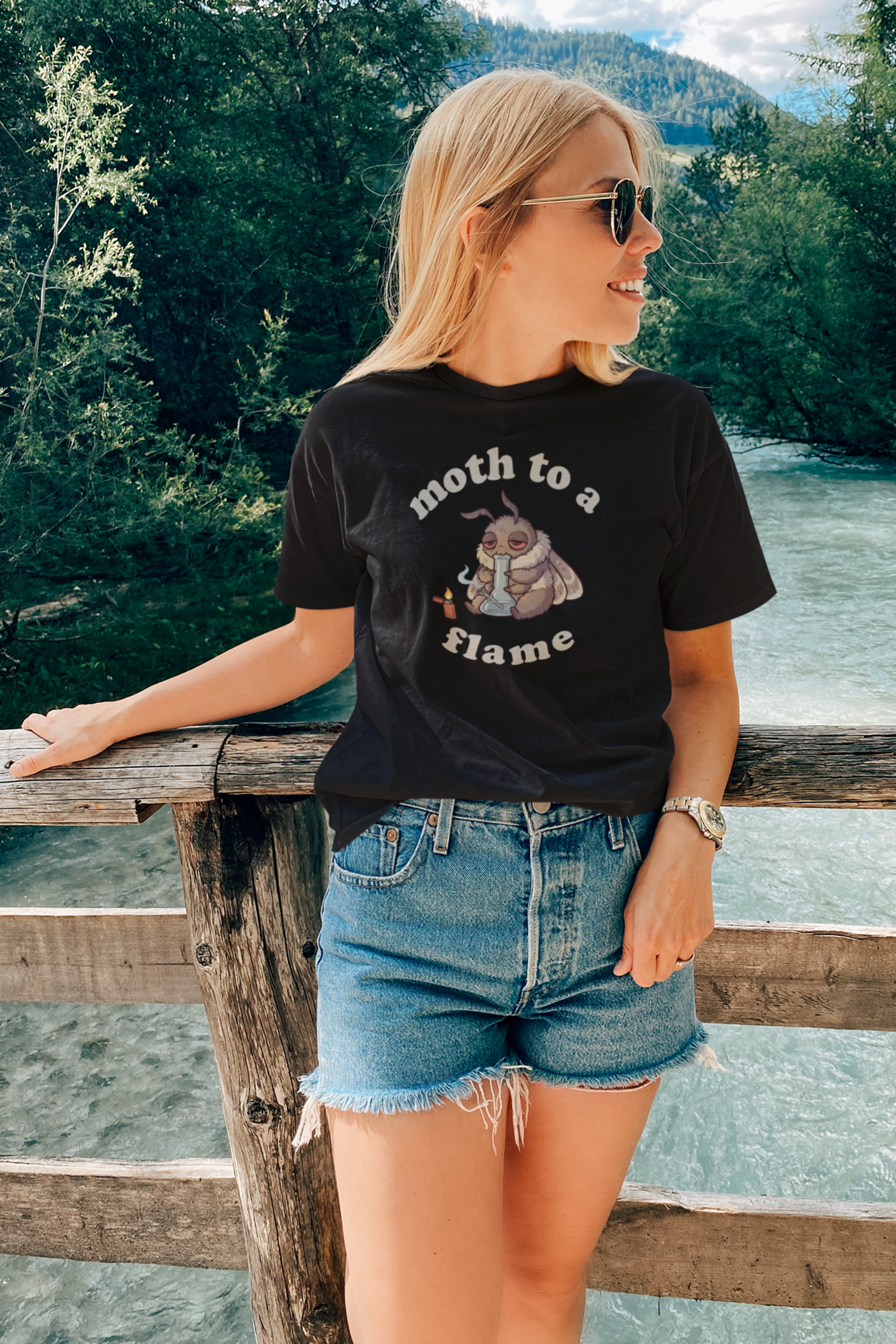 Woman wearing black Moth To a Flame cannabis tee outdoors by a river, featuring a cute stoner moth graphic for 420 lovers, weed smokers, and moth t-shirt fans.
