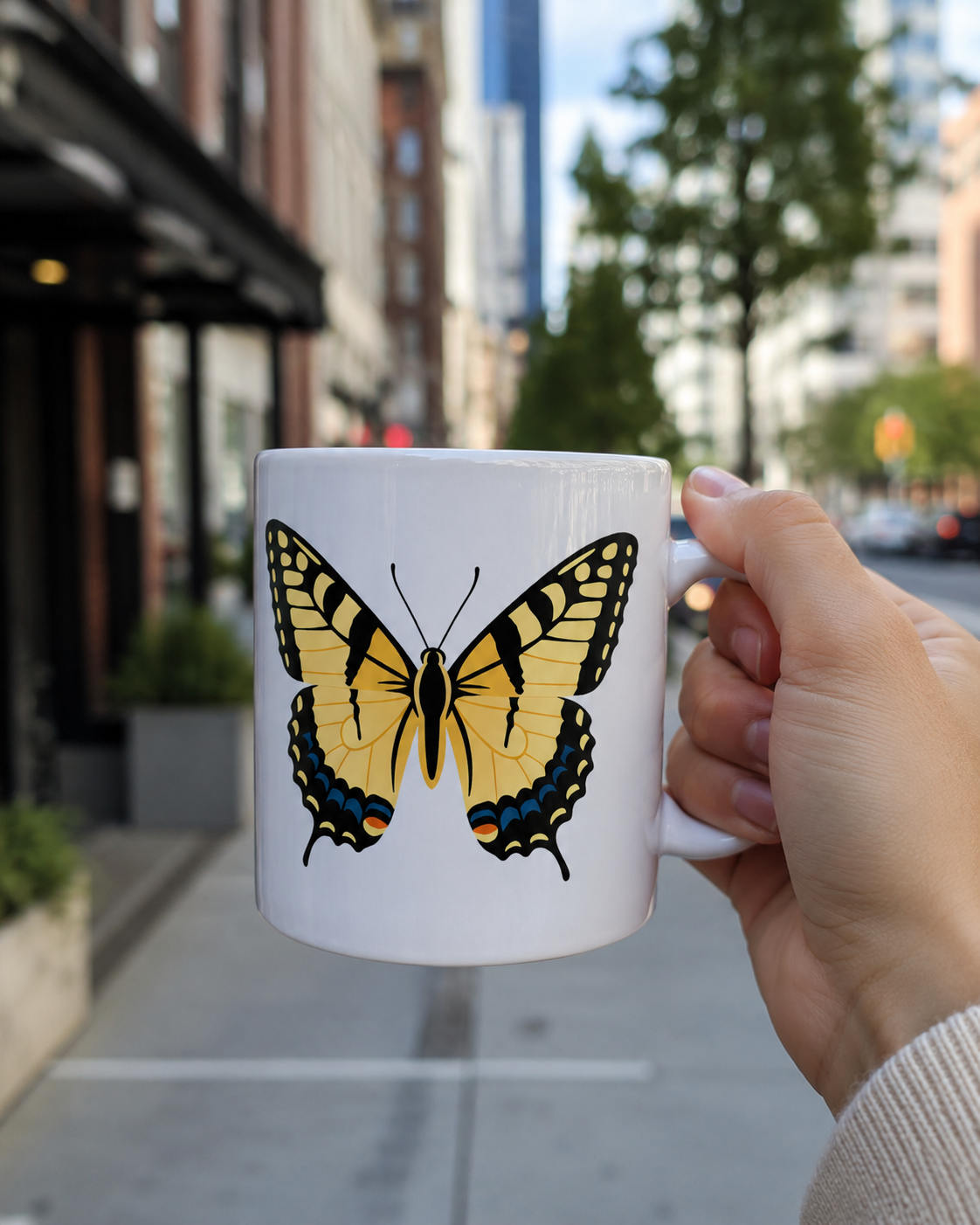 Hand holding a white butterfly coffee mug with a yellow and orange butterfly design in an urban outdoor setting.