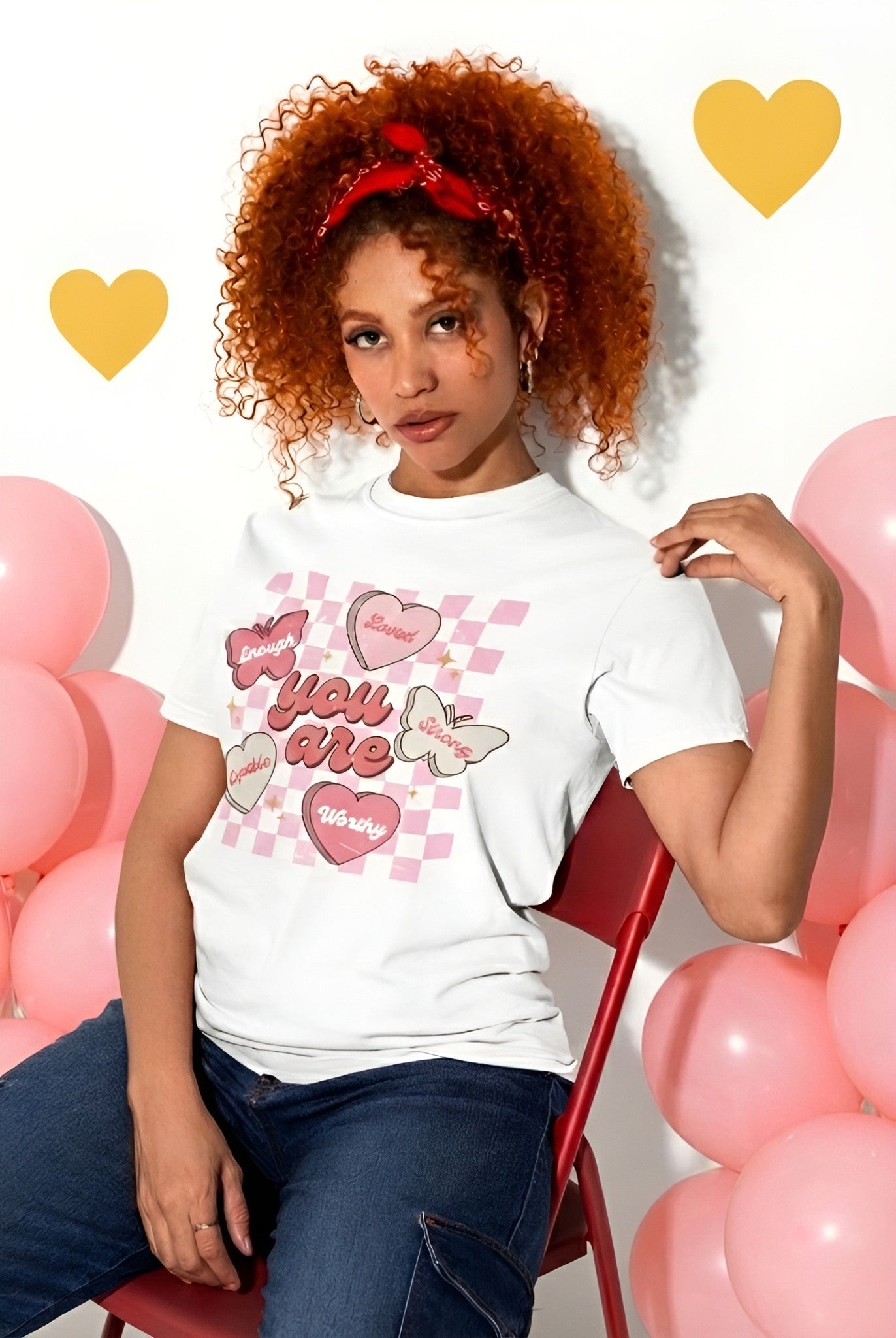 Woman wearing a white Valentine theme t-shirt with heart designs and text, sitting among pink balloons.